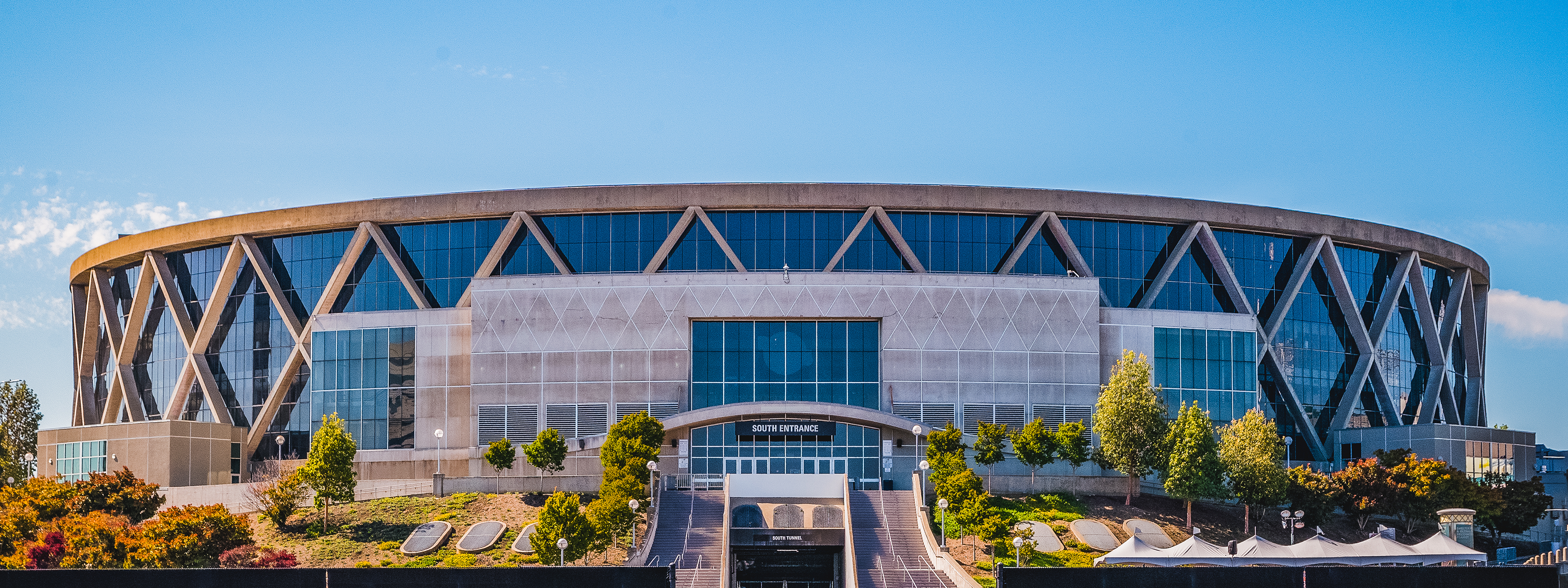 A wide view of a large, circular sports arena with geometric designs on the facade, large glass windows, and green trees in front under a clear blue sky.