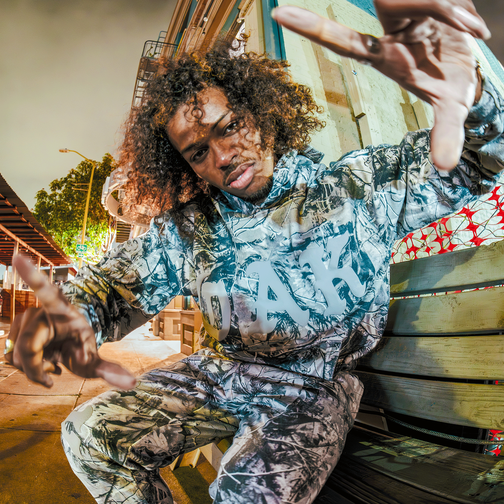 A person with curly hair wears an Oaklandish Cork Hoodie in grey camo, sitting on a wooden bench in a lit urban night scene, leaning forward with arms outstretched toward the camera.