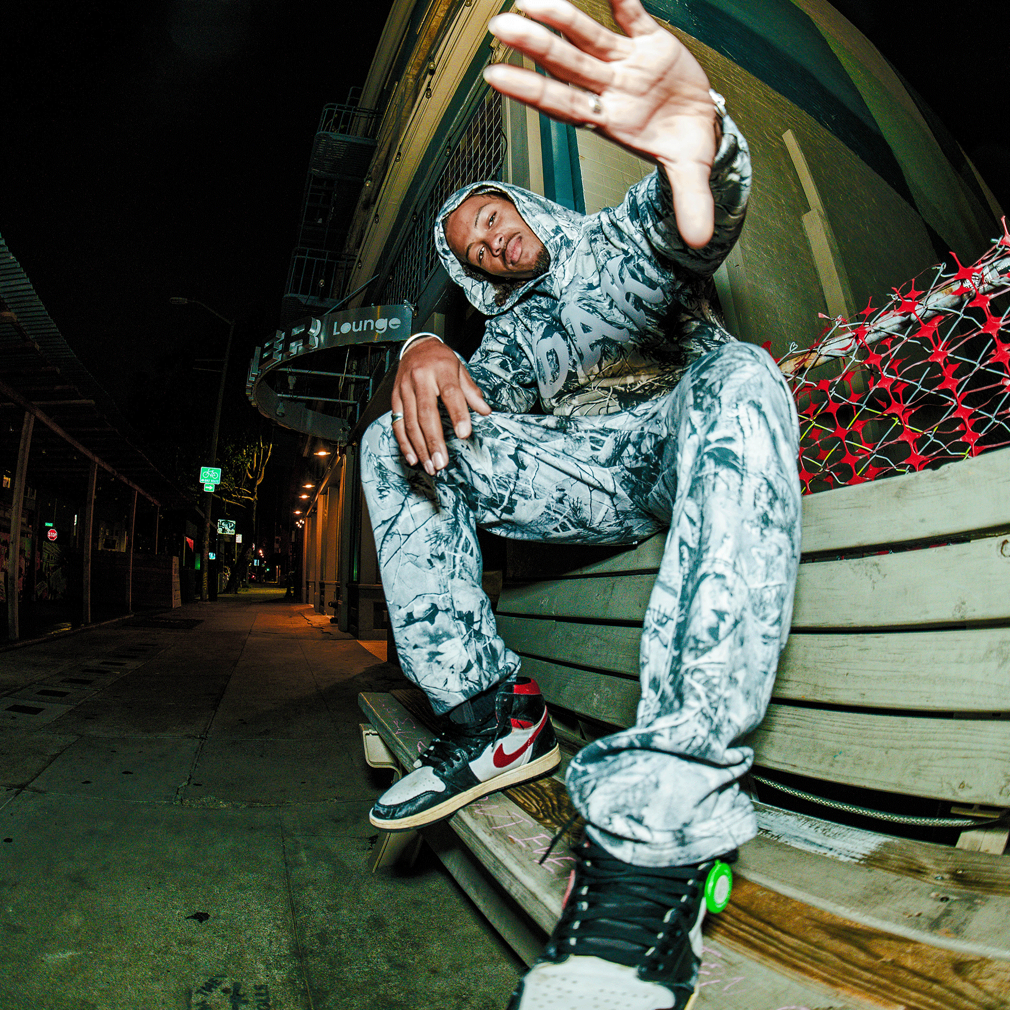 Wearing the Oaklandish Cork Hoodie with matching pants, a person sits on a night-lit urban bench, reaching toward the camera. Black, white, and red sneakers complete the look. A “lounge” sign glows in the city background.