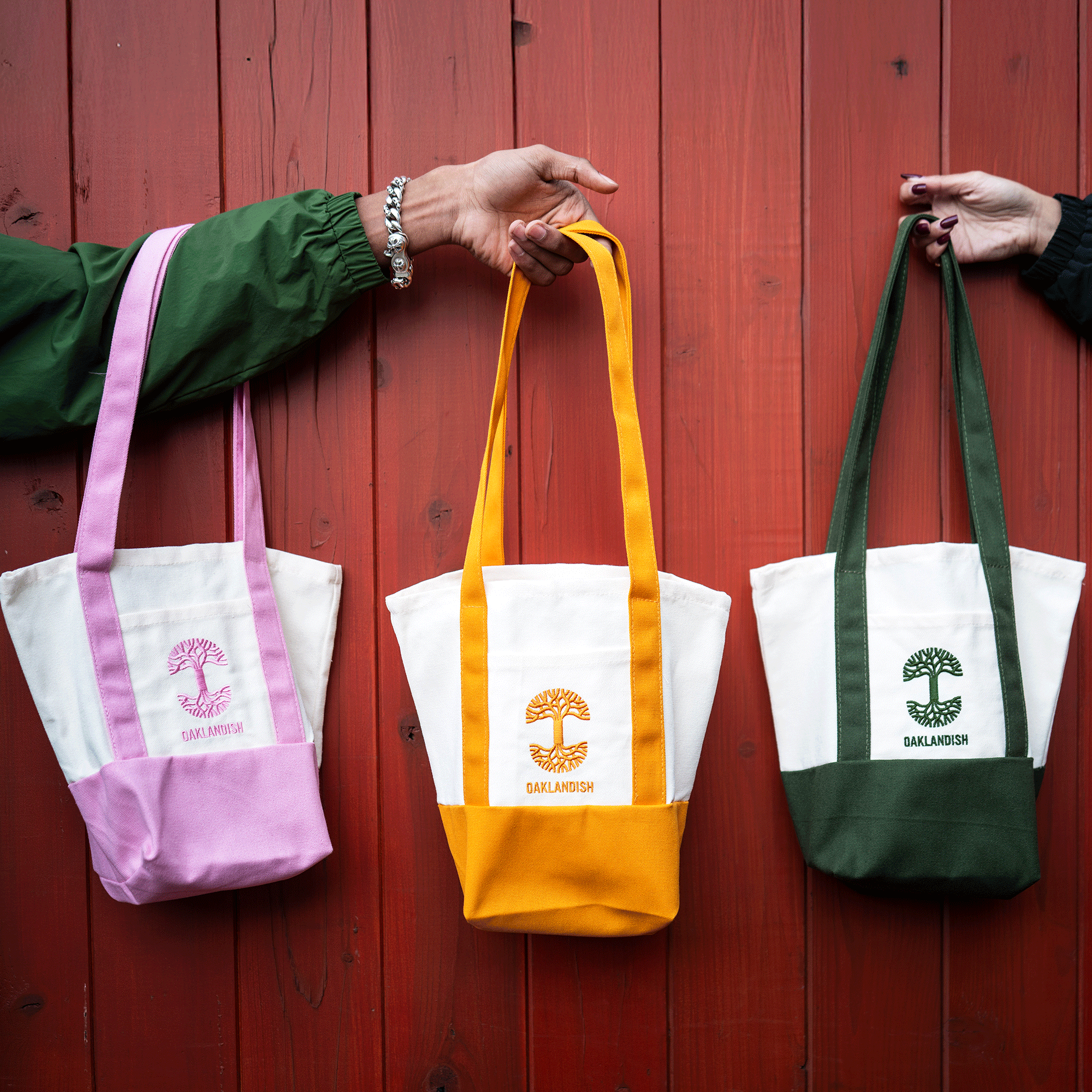 Two people hold up three Oaklandish Mini Grocery Totes by Oaklandish in front of a red wooden wall. The white canvas bags have long straps, colored accents in pink, yellow, and green, and display an embroidered tree logo and "OAKLANDISH" on the front.
