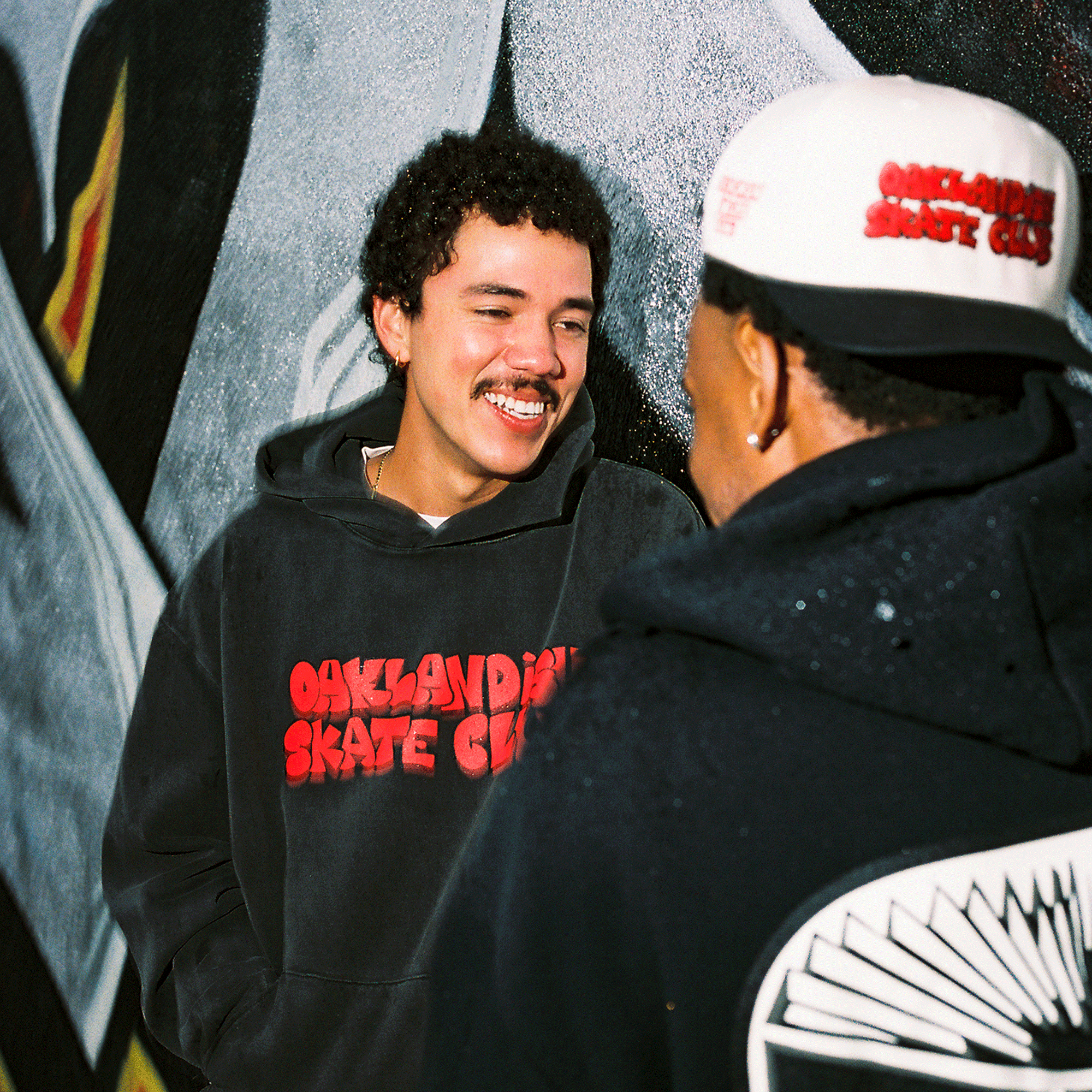 Two young men stand before a graffiti mural, smiling and chatting. Both wear black hoodies with "Oaklandish Skate Club" in red, while one rocks the Oaklandish Skate Club 2-Tone Hat, repping true skateboard culture.