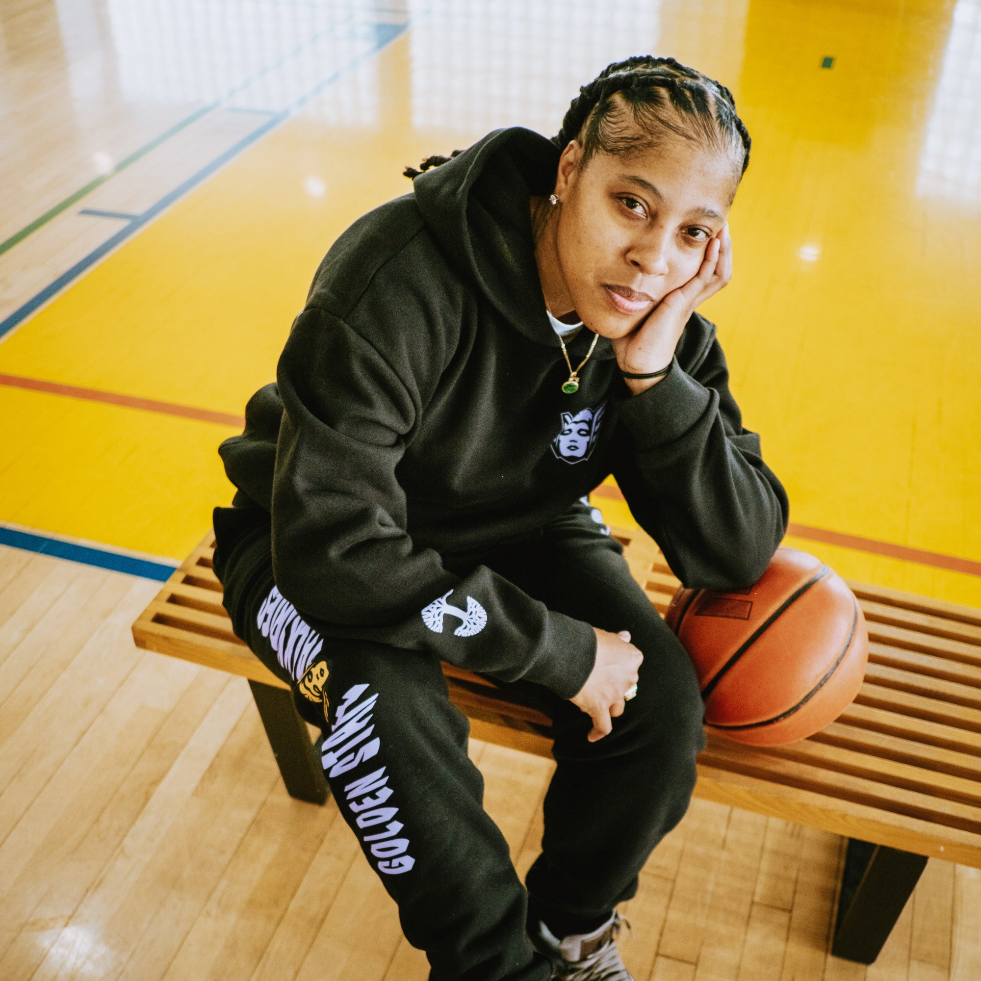 Wearing the GSV Renowned Pant and a black hoodie, a person sits on a wooden gym bench, resting their head on their hand beside a Golden State Valkyries basketball, with yellow paint highlighting the polished wood floor behind them.
