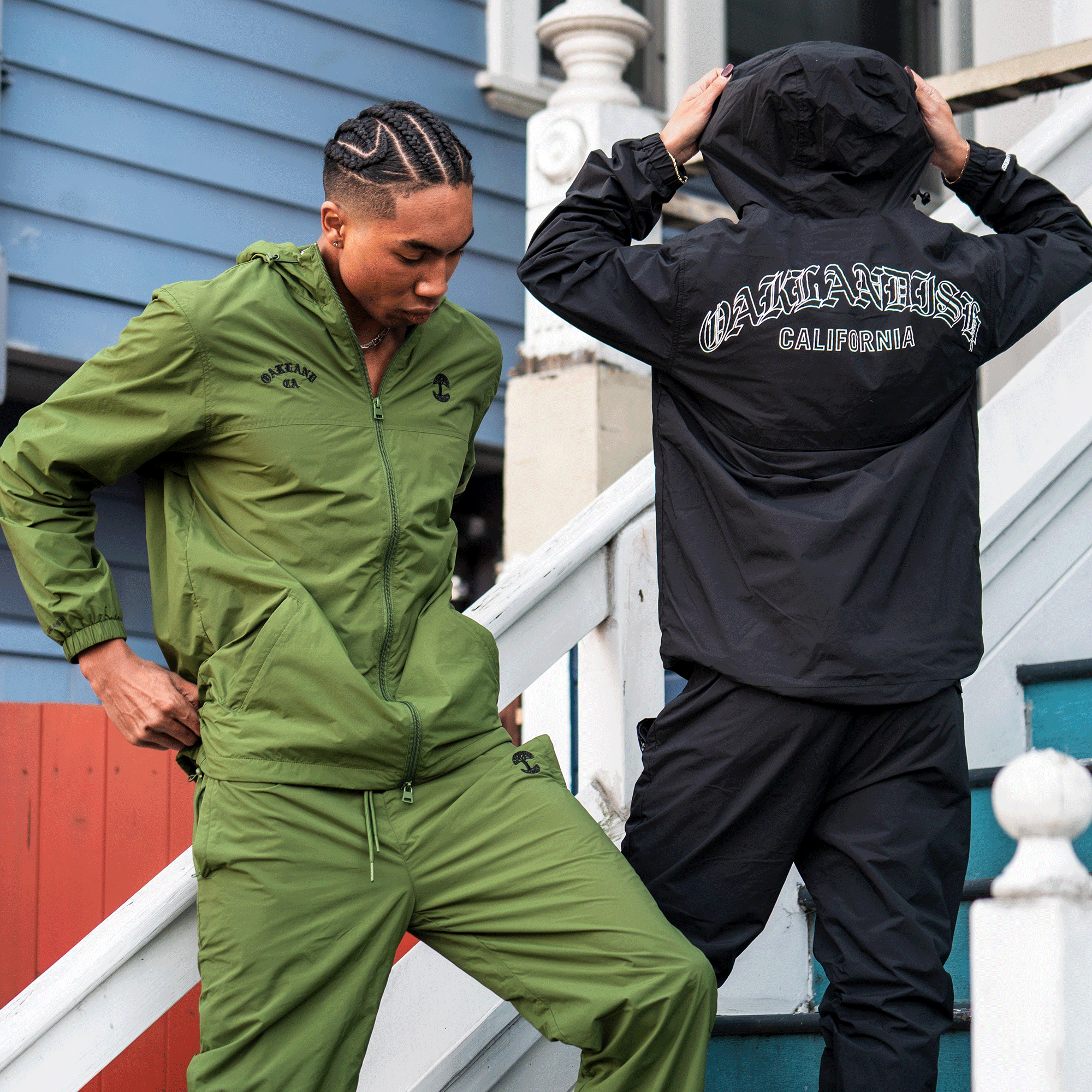 Two people stand on outdoor stairs in front of a blue house. One wears the Oaklandish Susurrus Track Top, while the other sports a men's hooded nylon zip jacket with "OAKLANDISH CALIFORNIA" printed on the back.