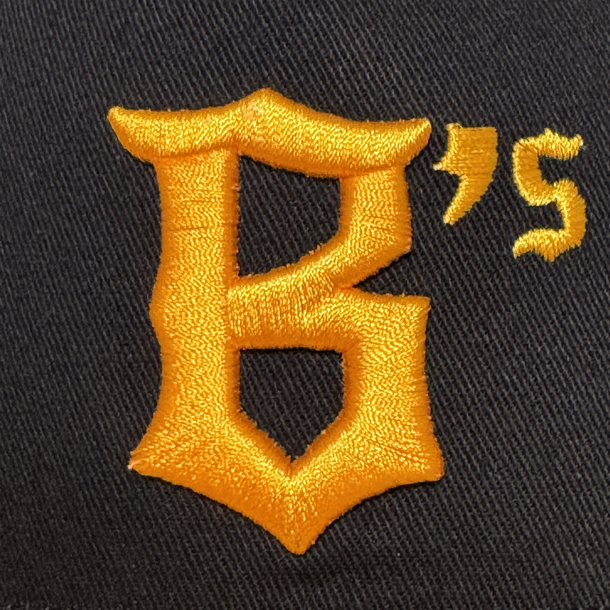 A close-up of black fabric on the Oakland Ballers 5 Panel Cap shows a large gold embroidered "B" in an ornate font, with a small gold "’s" above—detailed stitching pops, representing the Oakland Ballers brand.