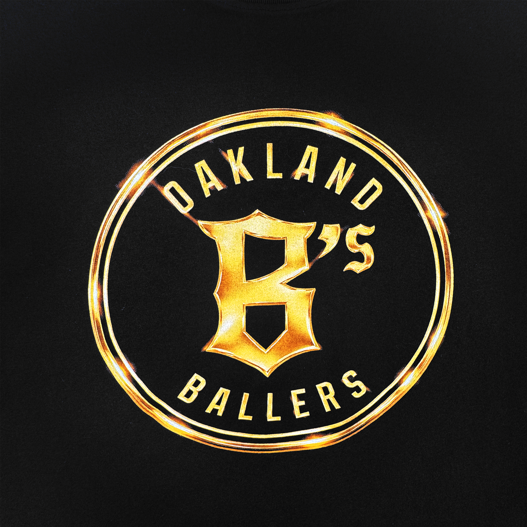 The Oakland Ballers Gold Tee features a circular gold emblem on black with "OAKLAND BALLERS" encircling a bold “B” and smaller "'S", highlighted by radiant metallic gold reflections. Brand: Oakland Ballers.