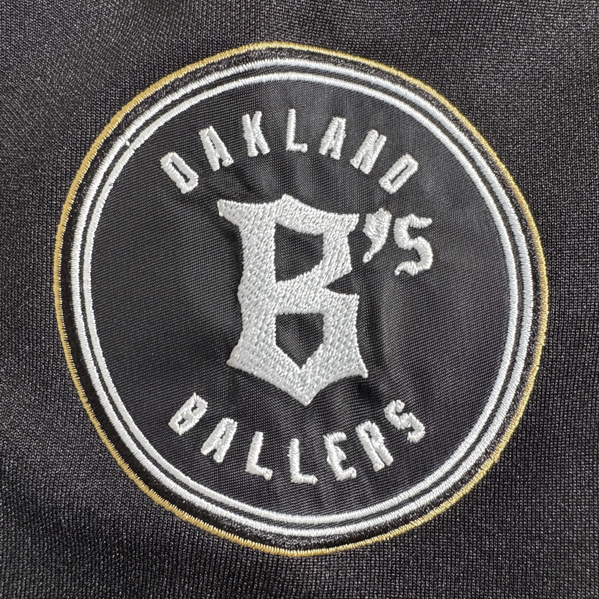 The Stands Up Jersey by Oakland Ballers features "Oakland Ballers" in white text encircling a large gold "B" and "'75" on a black background, bordered with white and gold rings.