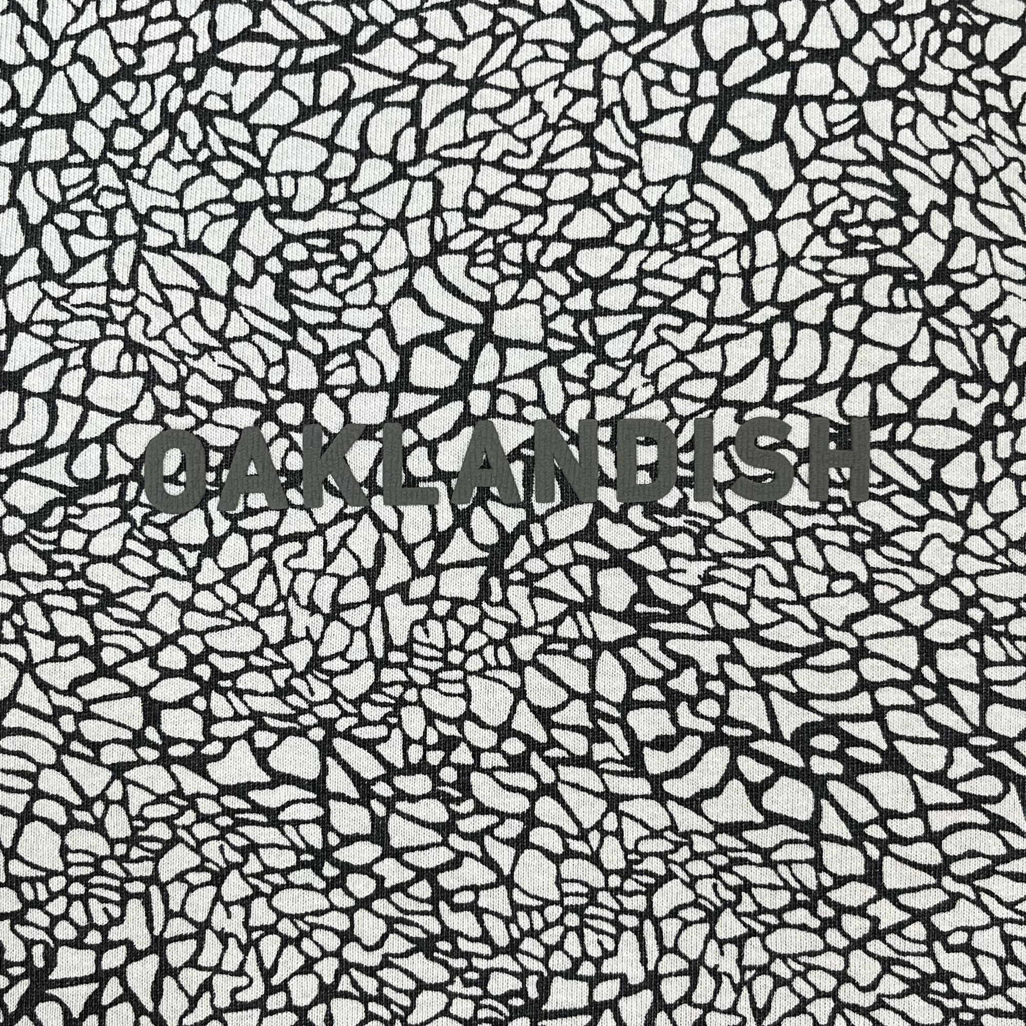 The image displays the Oaklandish Classic Logo Tee by Oaklandish, featuring the word “OAKLANDISH” in bold, dark letters centered on an abstract black and white crackle-patterned background reminiscent of tree bark or shattered glass. The intricate design is printed with a striking puff print technique that covers the entire shirt for a dynamic visual effect.