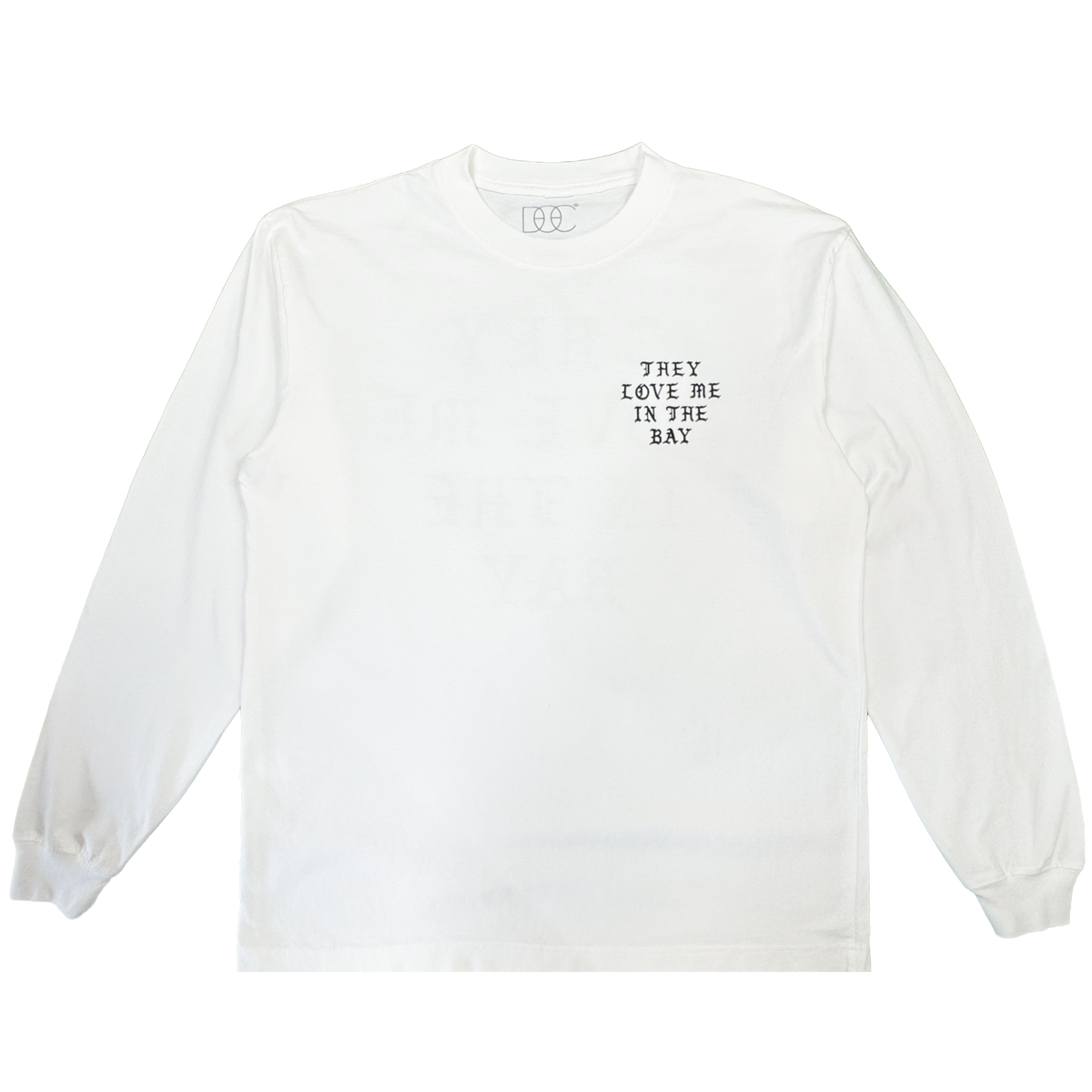 A heavyweight cotton long sleeve t-shirt by By DOC, the They Love Me LS Tee By Doc, is shown in men’s sizing laid flat against a plain white background. The only visible design is small black text on the left chest that reads, “THEY LOVE ME IN THE BAY.” There are no other embellishments or graphics.