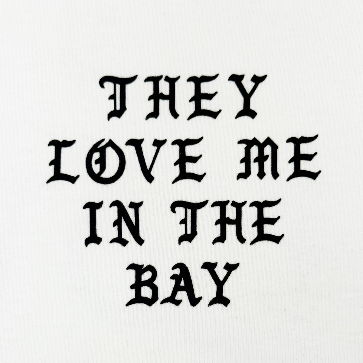 The They Love Me LS Tee By Doc by By DOC features bold, black gothic-style text that reads “THEY LOVE ME IN THE BAY,” centered and evenly spaced across three lines on a crisp white heavyweight cotton long sleeve t-shirt. Designed with men’s sizing, it combines standout style with exceptional comfort.