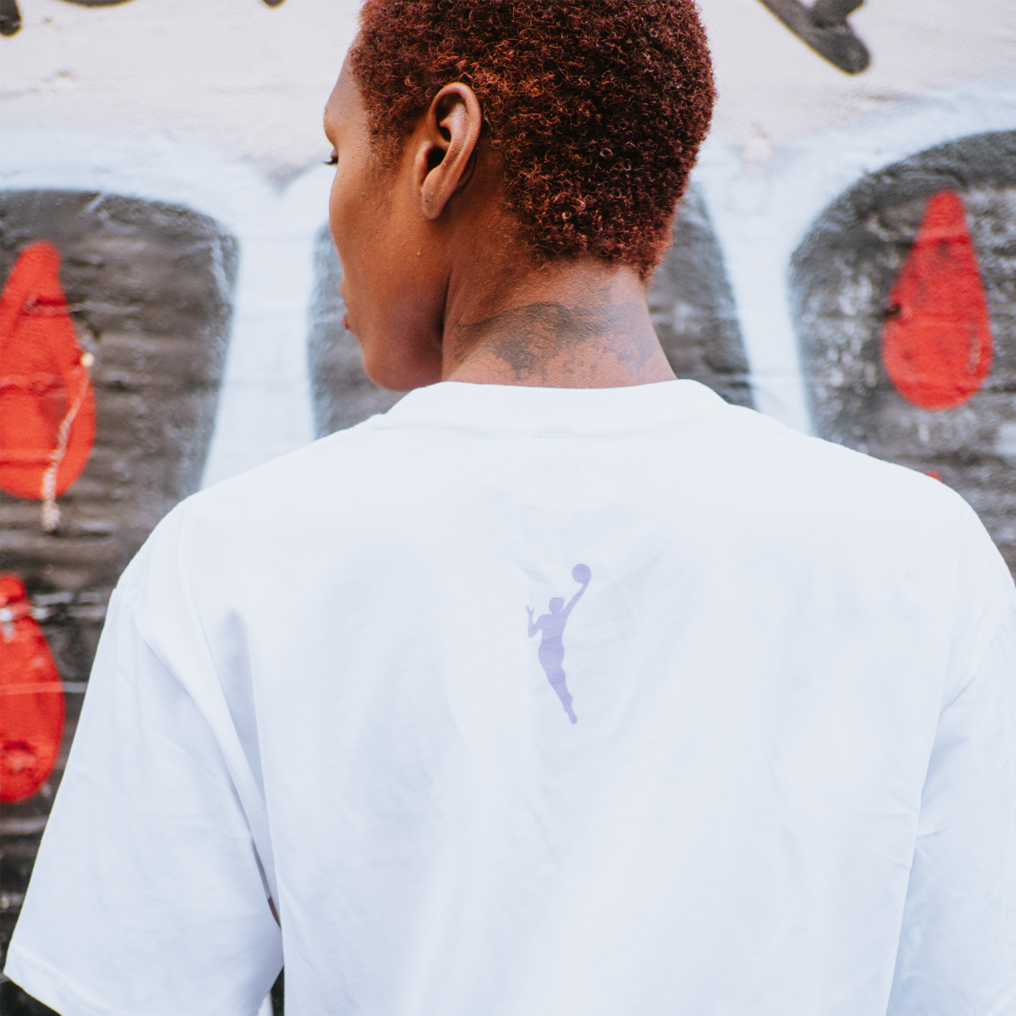 A person with short, reddish hair is seen from behind wearing the GSV Logo Love Tee by GSV—a 100% cotton classic fit shirt with a small basketball player silhouette logo, against a Golden State Valkyries-style backdrop.