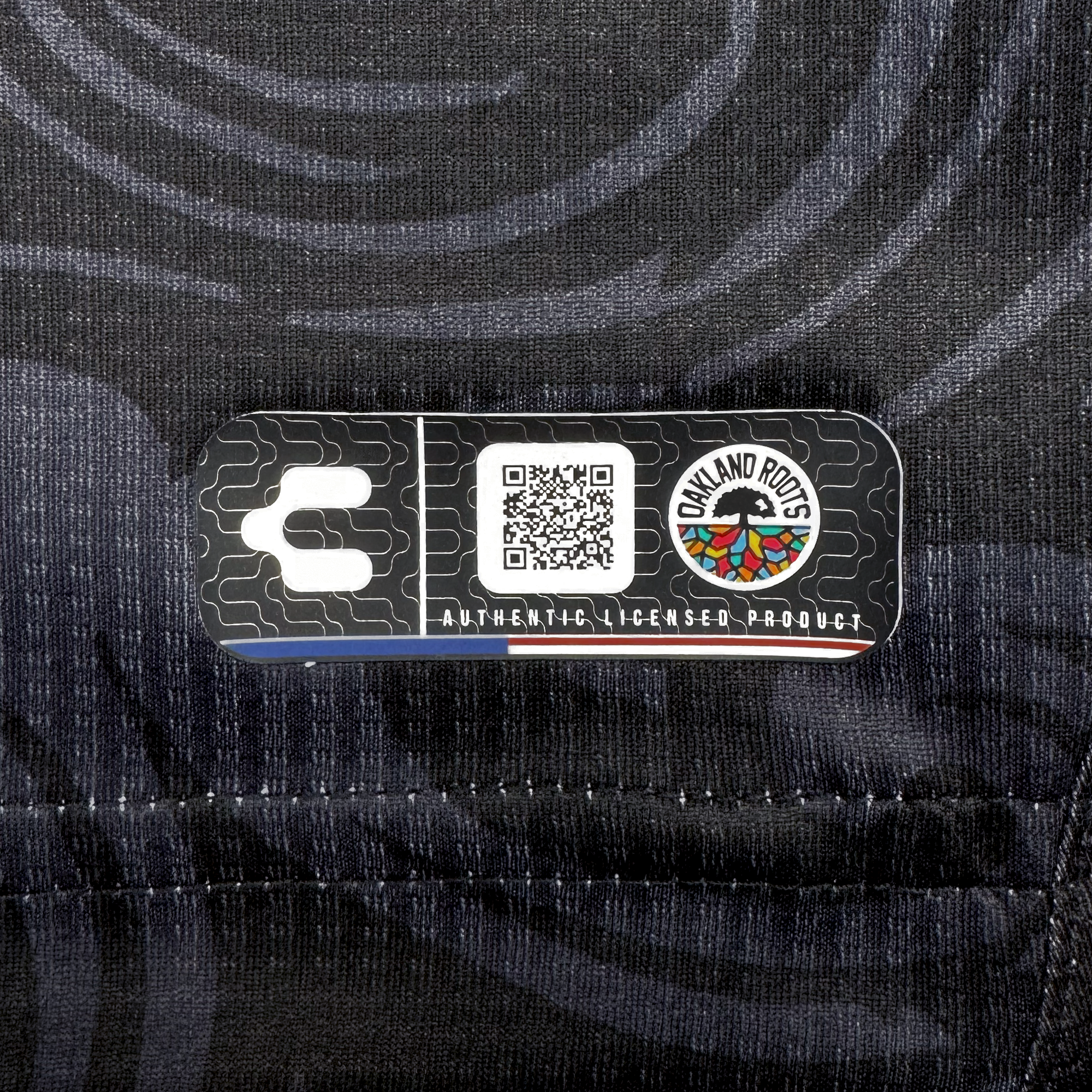 Close-up of the Unisex Oak-ness Awakens Kit’s black fabric tag, featuring Oakland Roots SC’s white “C” logo, a QR code, and an Oak-ness Monster-inspired logo. Below, “AUTHENTIC LICENSED PRODUCT” appears in white text.