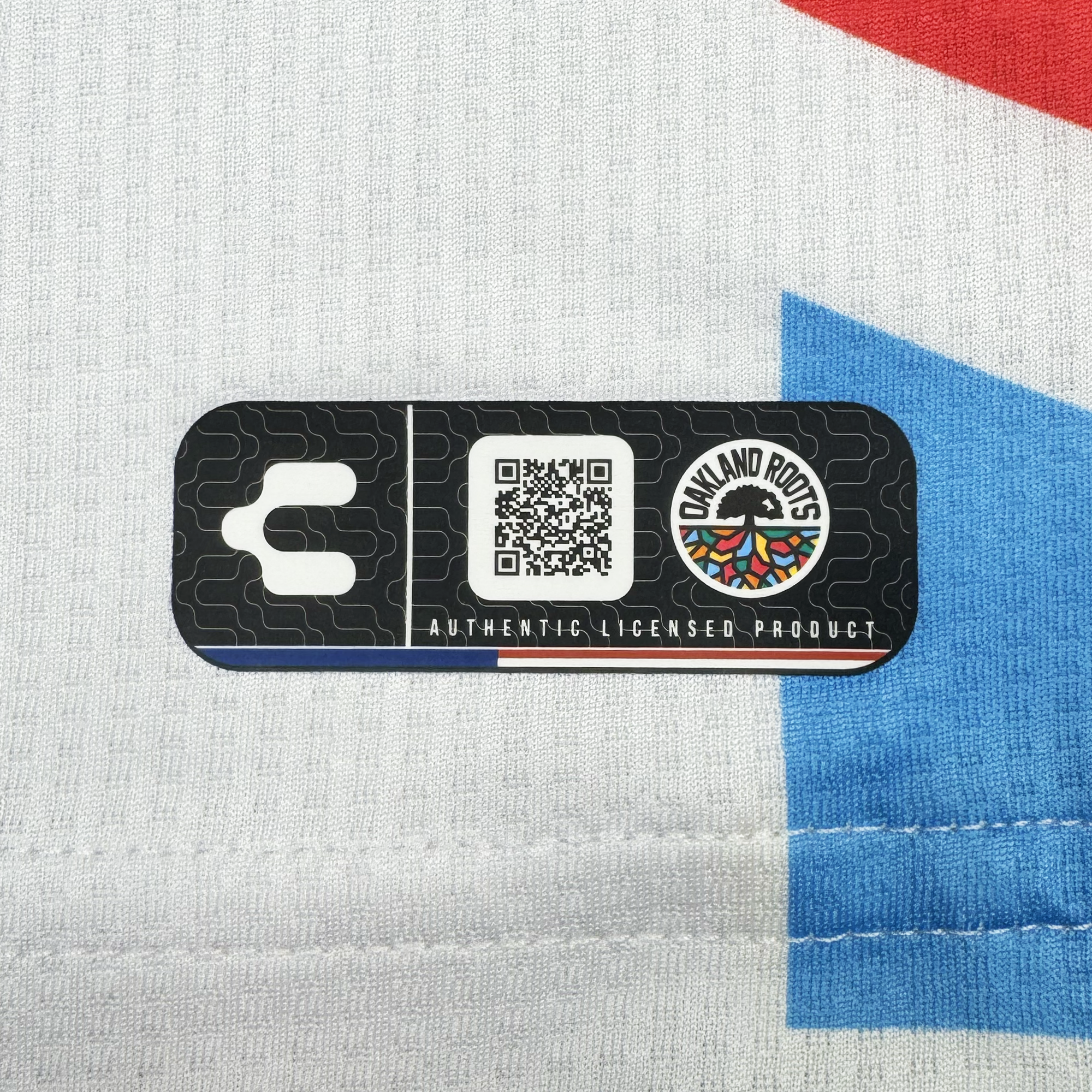 A black rectangular label is stitched onto the Unisex Mosaic in Motion Kit by Oakland Roots SC, featuring a QR code, plug icon, the club logo, and "AUTHENTIC LICENSED PRODUCT" text on a white fabric with red and blue mosaic patterns.