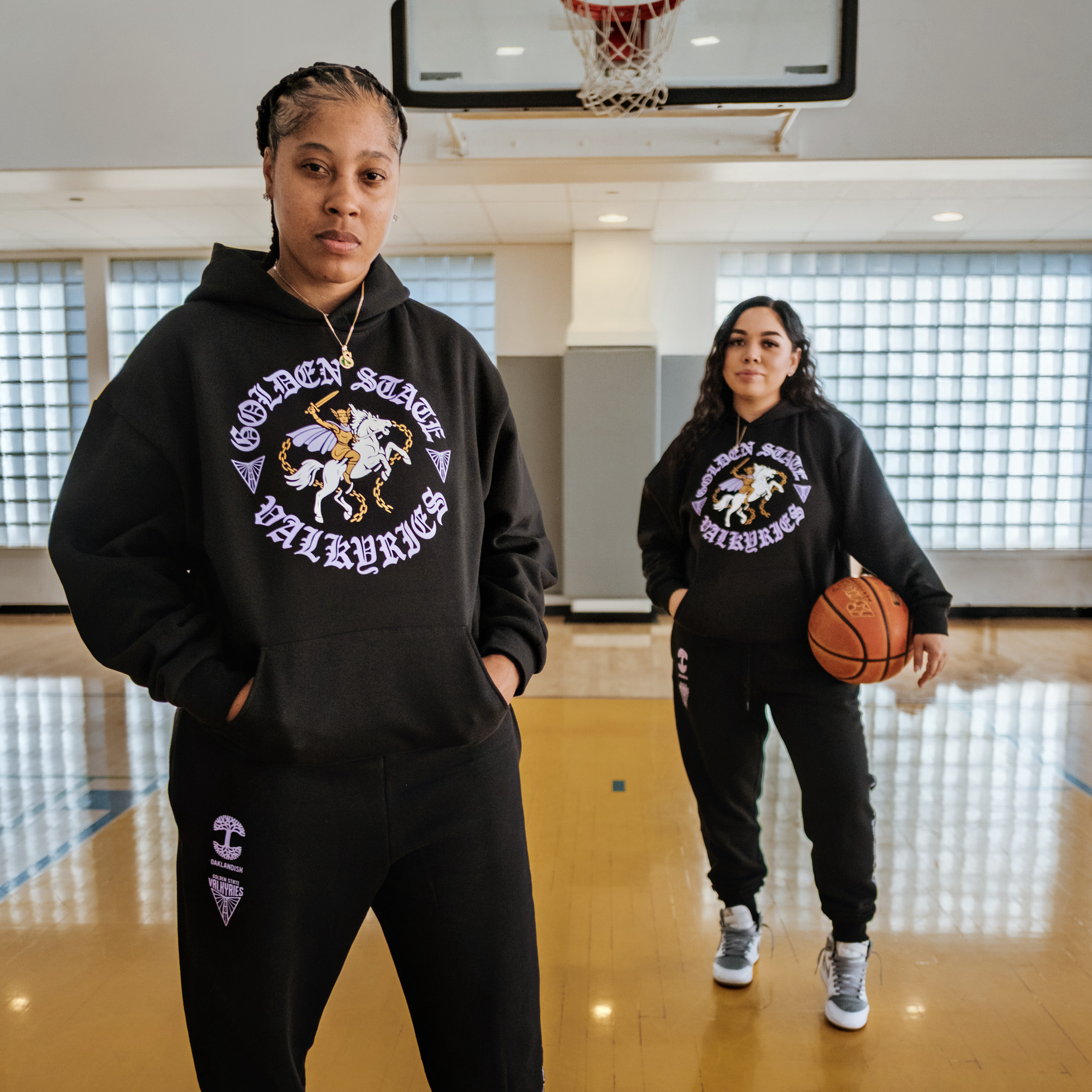 Two women stand on an indoor basketball court, both in black "Golden State Valkyries" hoodies featuring a valkyrie on horseback. One holds a basketball. Their hoodies are men's sizing, styled with GSV ROTV Pant sweatpants from GSV.