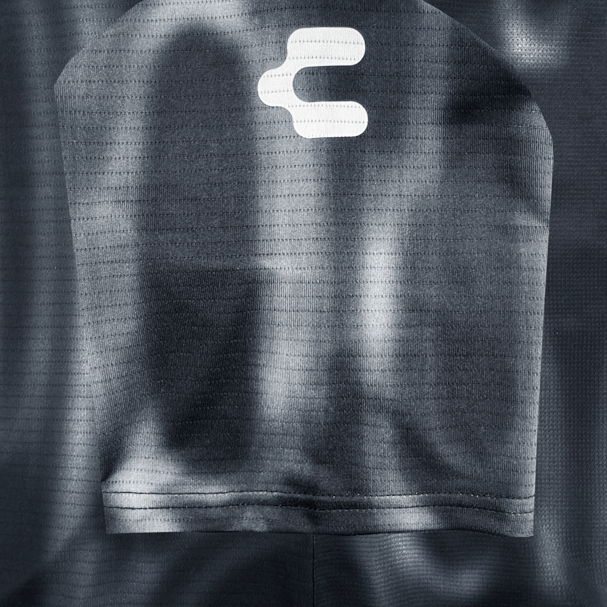 A close-up of the dark, textured polyester fabric of the 2026 OAKLAND SOUL SC SPIRIT JERSEY shows a curved seam with stitched edge and subtle shadows, featuring a bold white Oakland Soul logo near the top.