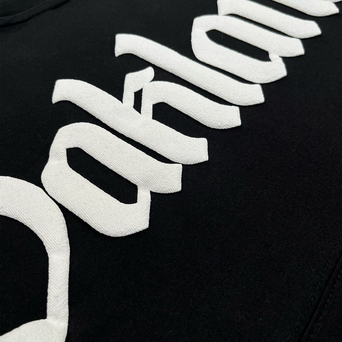 Close-up of black fabric with raised, white, textured letters spelling “Oakland” in a bold Old English-style font. The stitching detail is visible, emphasizing the embroidered texture against the smooth background of this 100% cotton men’s Temple LS Tee by Oaklandish.