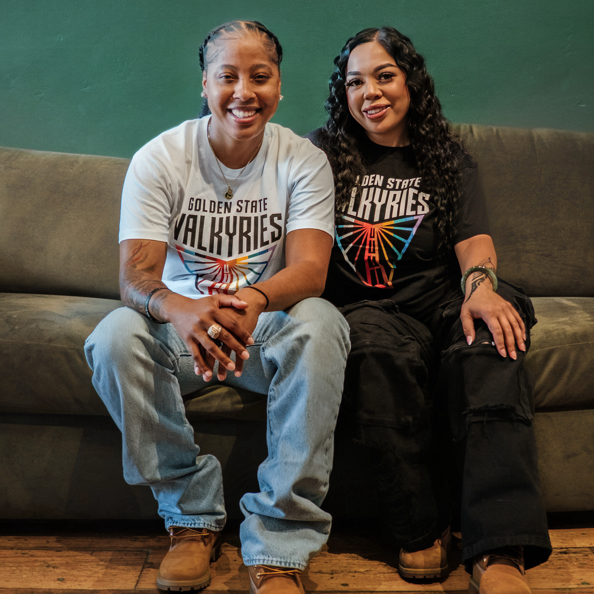 Two people sit on a gray couch against a green wall, both wearing classic fit GSV Logo Love Tees—one white, one black—by GSV. Made from 100% cotton, the tees pair perfectly with jeans and tan boots for a comfortable, stylish look.