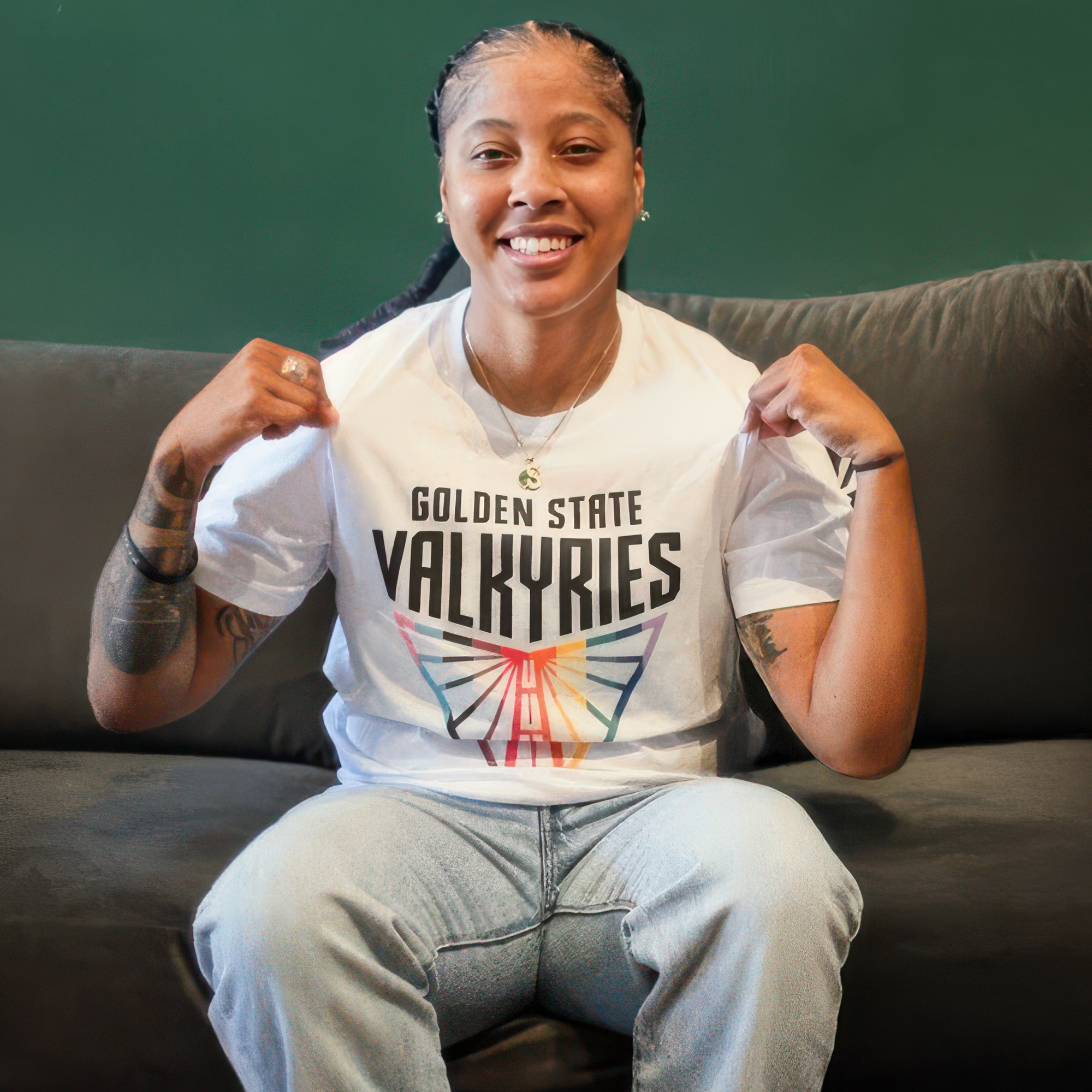 A person with braided hair smiles on a gray couch, showing off their GSV Logo Love Tee by GSV, featuring a colorful logo. They wear light blue jeans, display arm tattoos, and sit against a green wall.