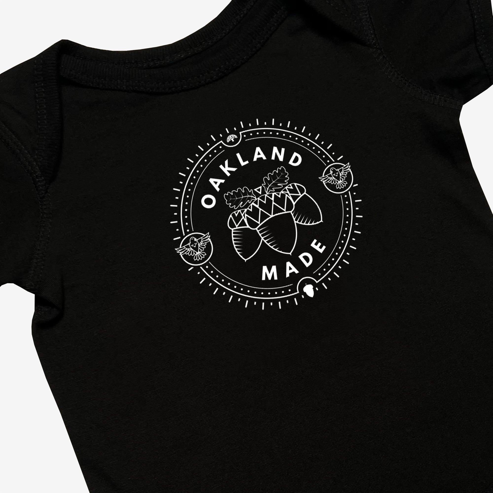 Close-up of black infant one-piece with a white circle graphic with the words “Oakland Made” and hand-drawn chestnuts.