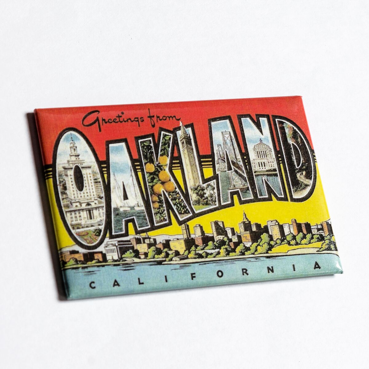 The "Greetings from Oakland Magnet" by Oaklandish is a versatile and unique refrigerator magnet featuring the bold word "OAKLAND" filled with colorful illustrations of local landmarks and scenes. The background transitions from red at the top to yellow, with a blue strip at the bottom, making it not just functional but also aesthetically pleasing.