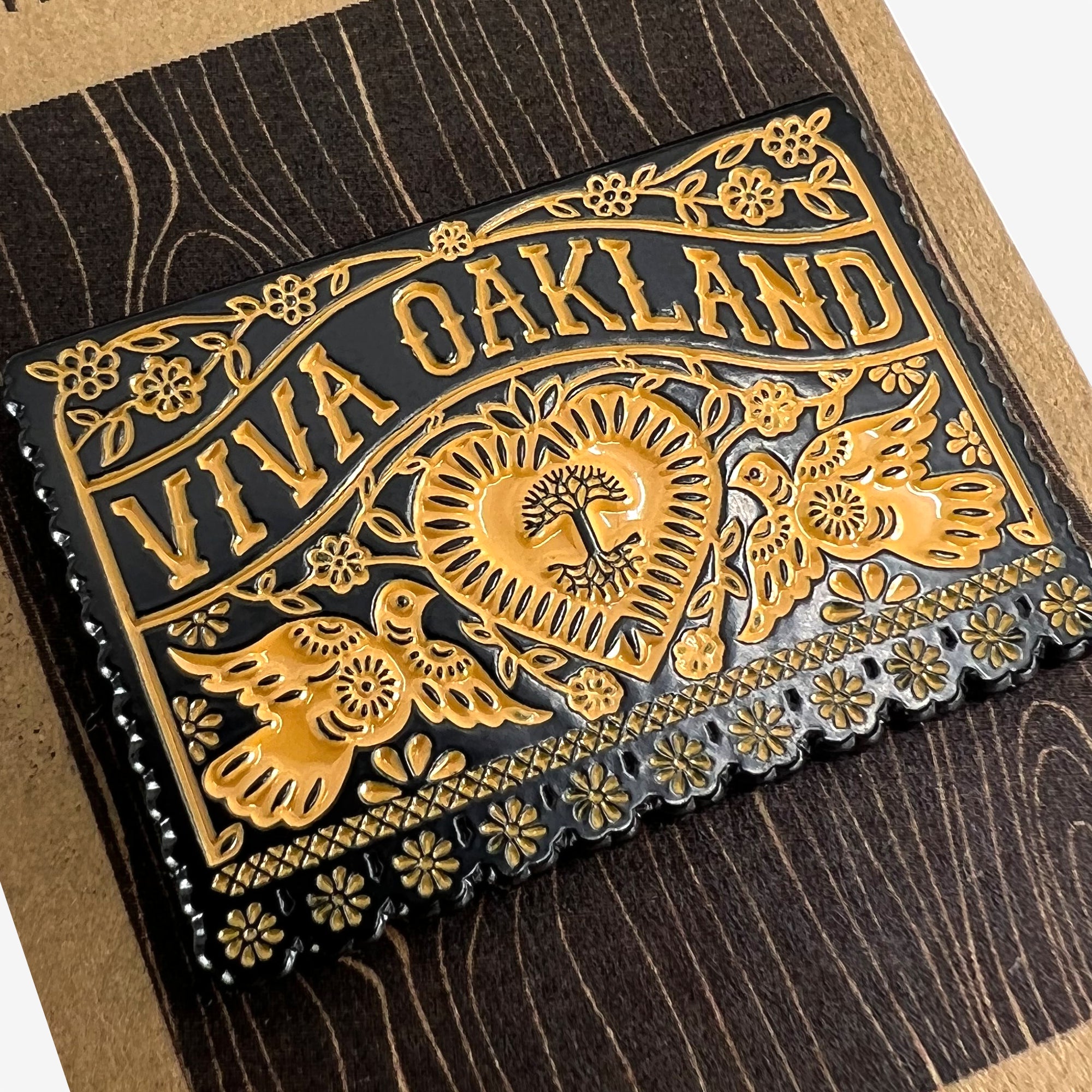 Close-up of gold and black enamel pin Viva Oakland wordmark with heart, birds and flowers on brown paper retail packaging.