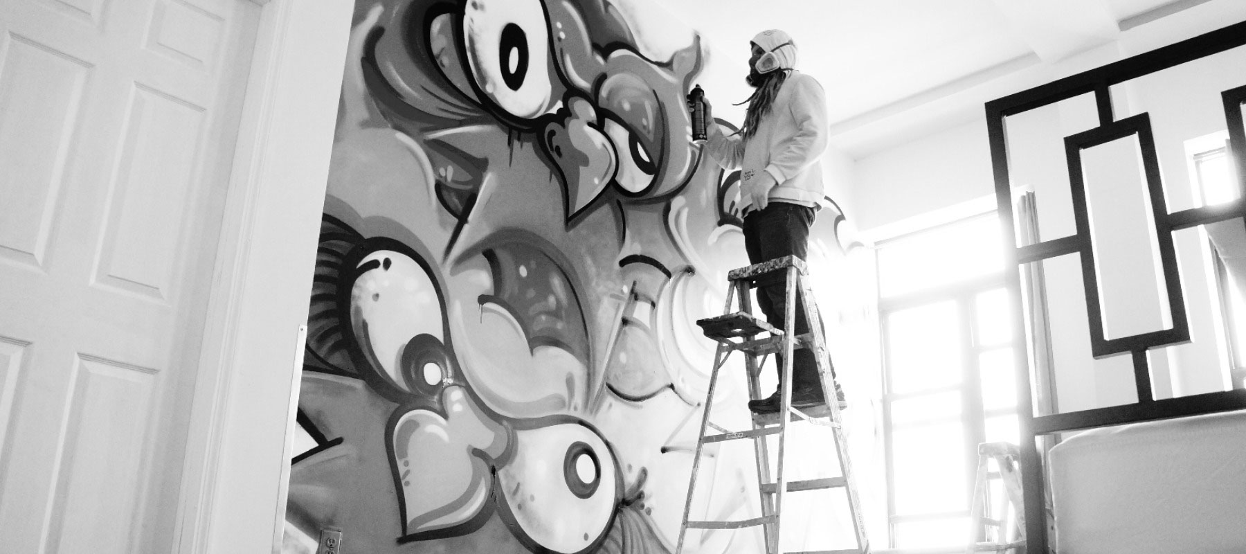 A person in a hoodie and mask stands on a ladder, spray-painting a large, colorful graffiti mural with abstract eyes and shapes on an indoor wall; sunlight streams through nearby windows.