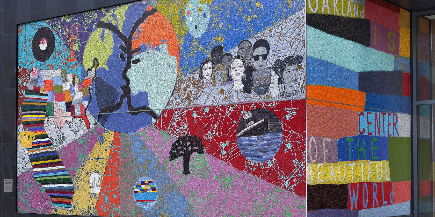 A colorful mural features a globe, diverse faces, a tree, abstract shapes, and text reading “OAKLAND IS… CENTER OF THE BEAUTIFUL WORLD,” symbolizing diversity, unity, and community in Oakland.