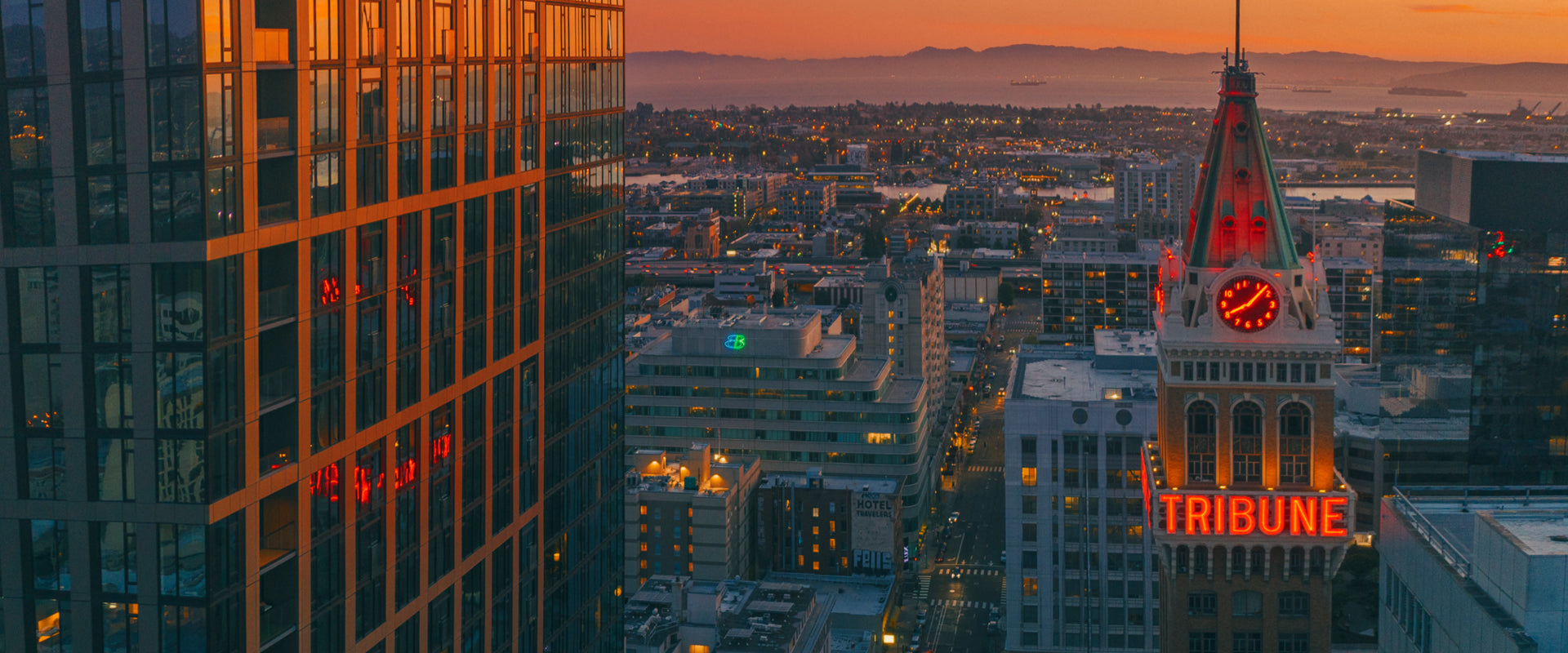 A cityscape at sunset features the Tribune Tower with its red neon sign and clock, modern glass buildings, and distant hills under an orange sky, reflecting the glow of the setting sun.