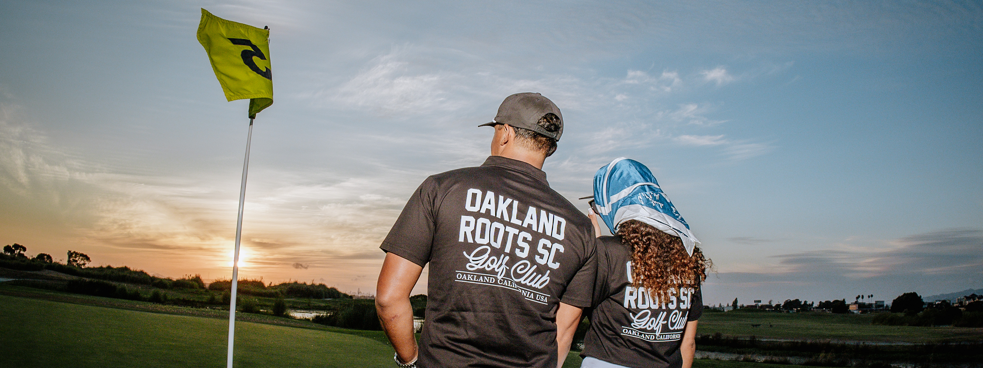 Two people wearing "OAKLAND ROOTS SC Golf Club" shirts stand near a golf flag on a green at sunset, looking towards the horizon; one wears a cap, the other has a scarf on their head.