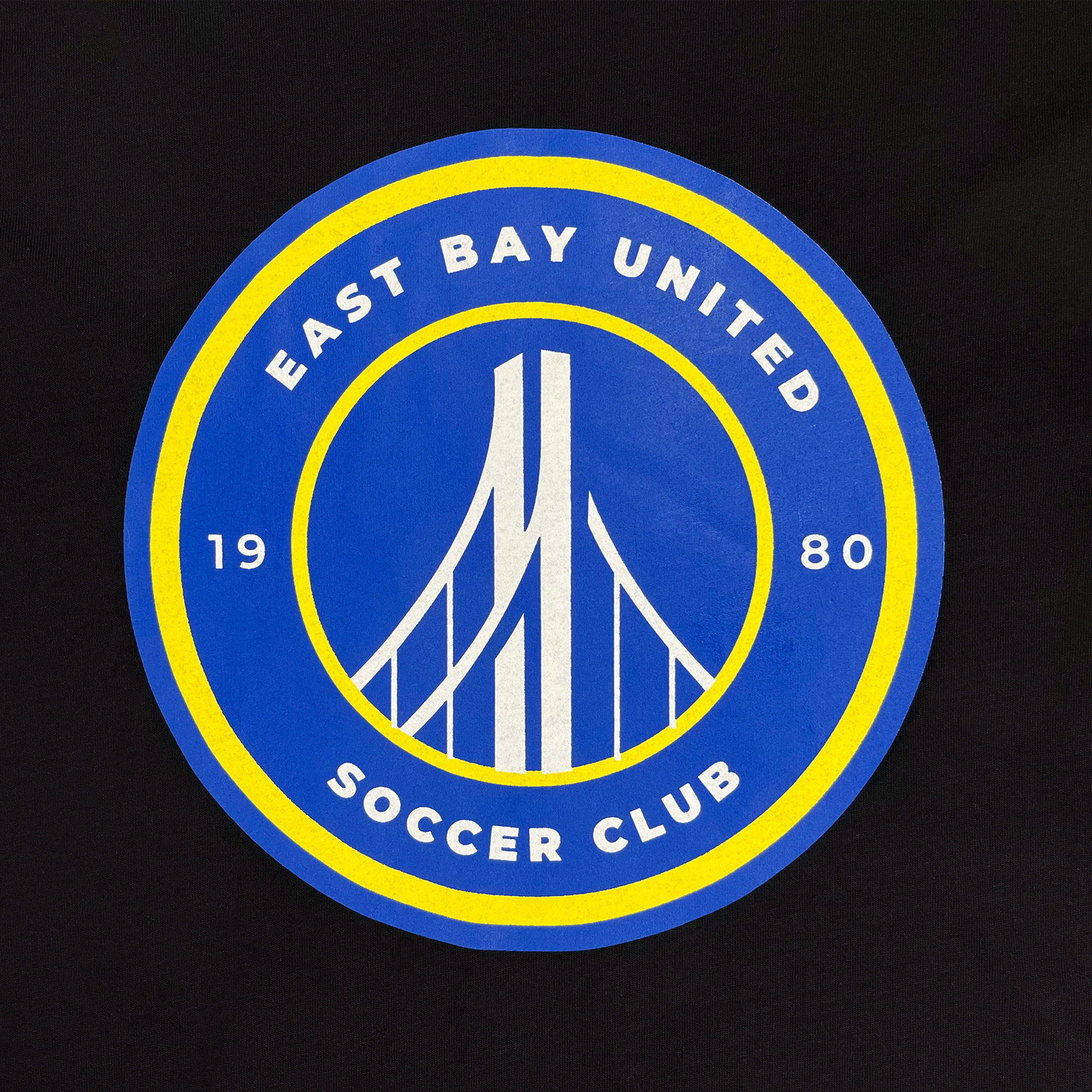 The EBU Long Sleeve Tee from Supported Brands showcases a striking logo for the East Bay United Soccer Club, a dedicated youth soccer club. The design features a stylized bridge in white set against a blue background. "East Bay United" is prominently displayed at the top of the blue circle, with "Soccer Club" at the bottom and "1980" split on either side in yellow. A yellow border surrounds the entire design, completing this eye-catching look.