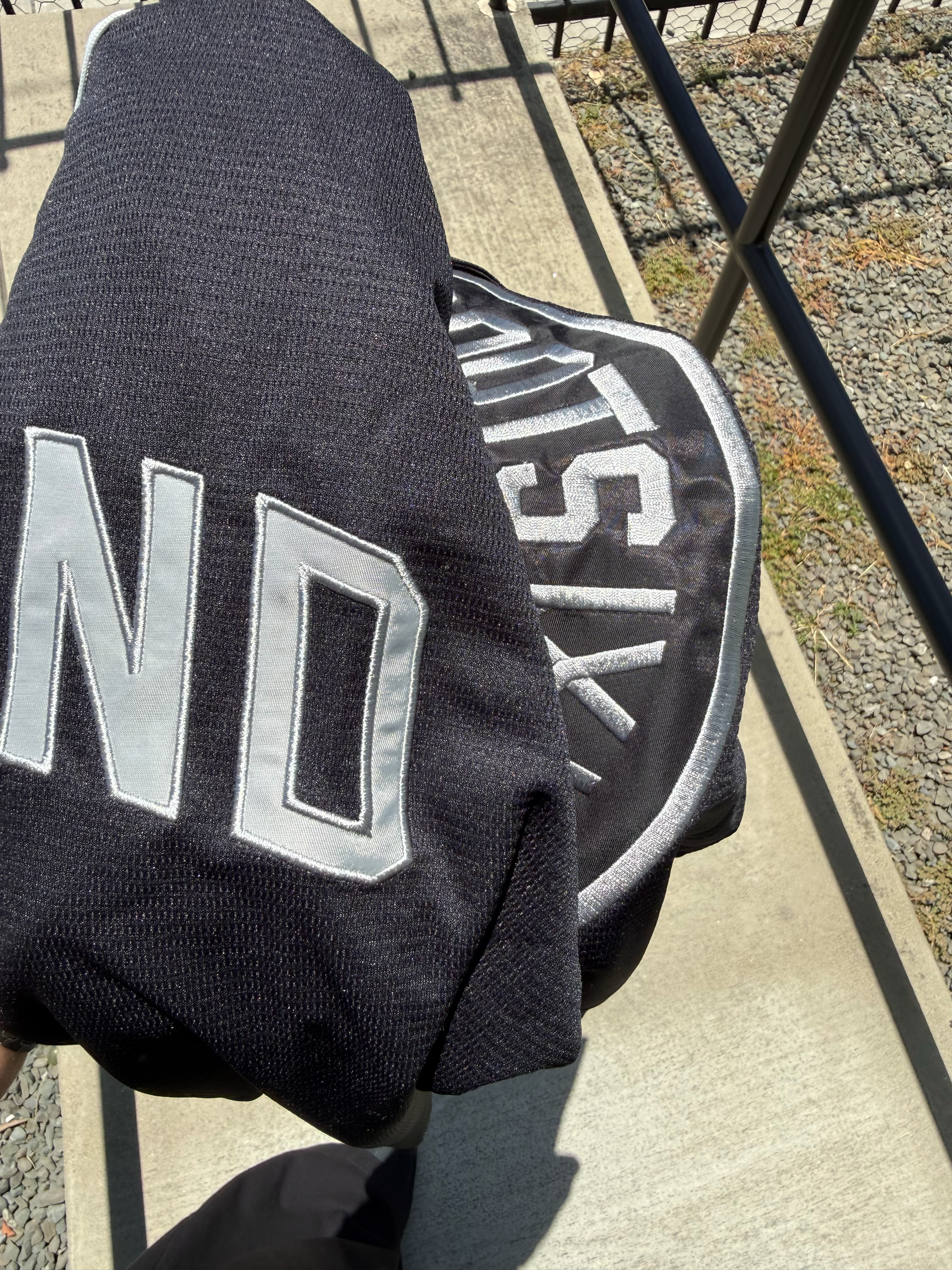 A person holds a folded ORSC Button Up Jersey by Oakland Roots SC, featuring bold white lettering and a partial circular logo, while standing on a sunlit concrete path beside a metal rail and gravel.