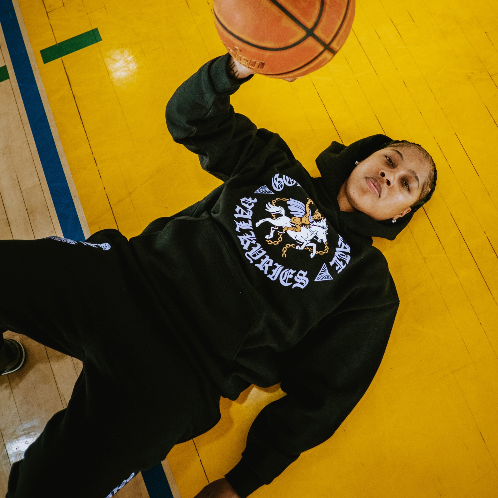 Wearing the GSV ROTV Hoodie by GSV, a person with braided hair and a focused gaze lies on a yellow basketball court in sweatpants, holding a basketball overhead. The black hoodie features bold “Golden State Valkyries” graphic text.