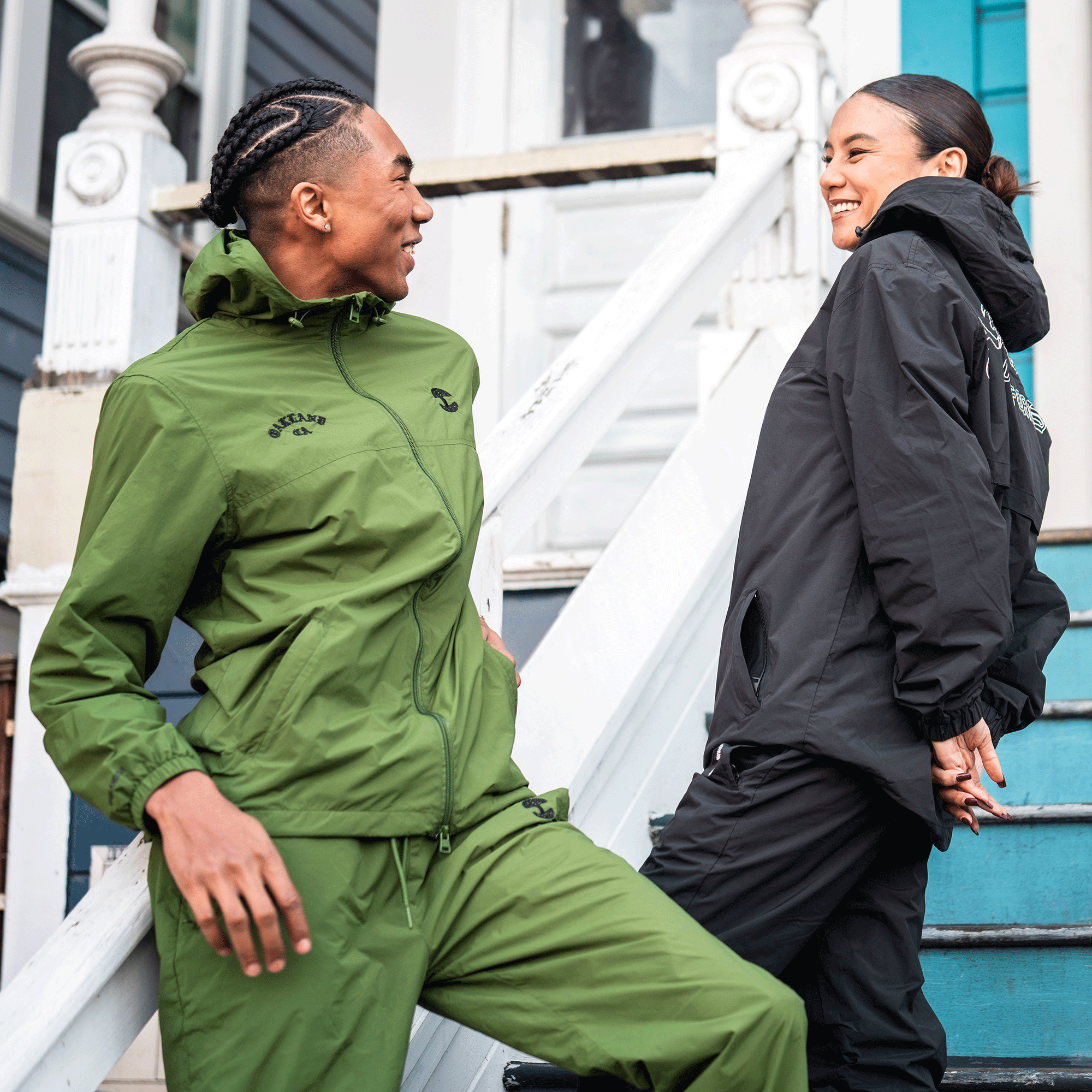 Two people smile at each other on a porch. The person on the left wears green nylon pants with an adjustable waistband. The person on the right stands by a white door in Oaklandish Susurrus Track Bottoms, featuring an embroidered tree logo.