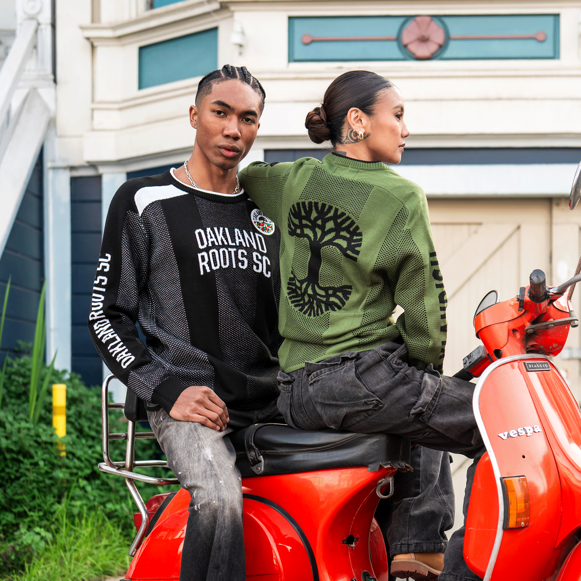 Two stylish young adults sit on a red Vespa. The man wears an Oakland Roots SC Clubhouse Sweater (unisex) by Oakland Roots SC, while the woman sports an Oaklandish knit sweater with a large tree on the back. A cozy house and lush plants complete the scene.
