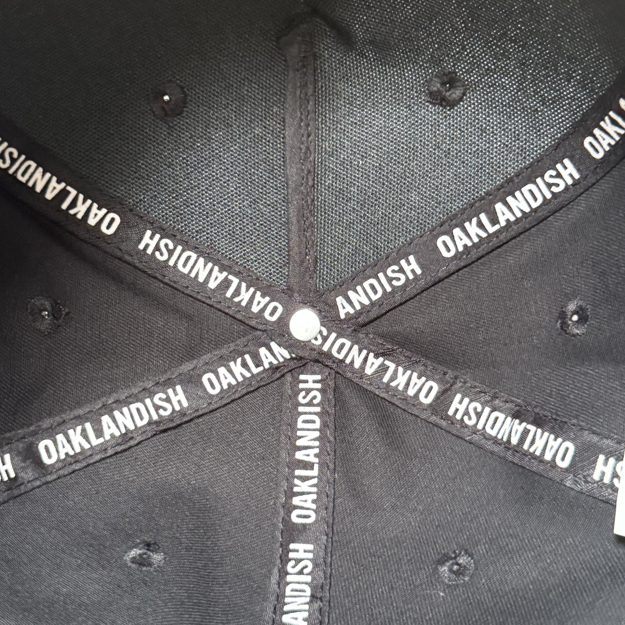 Close-up of the inside of the Oakland Ballers 5 Panel Cap. Black taped seams repeatedly feature "OAKLANDISH" in white, subtly referencing the Oakland Ballers baseball team. A small silver rivet is centered inside the black snapback hat.