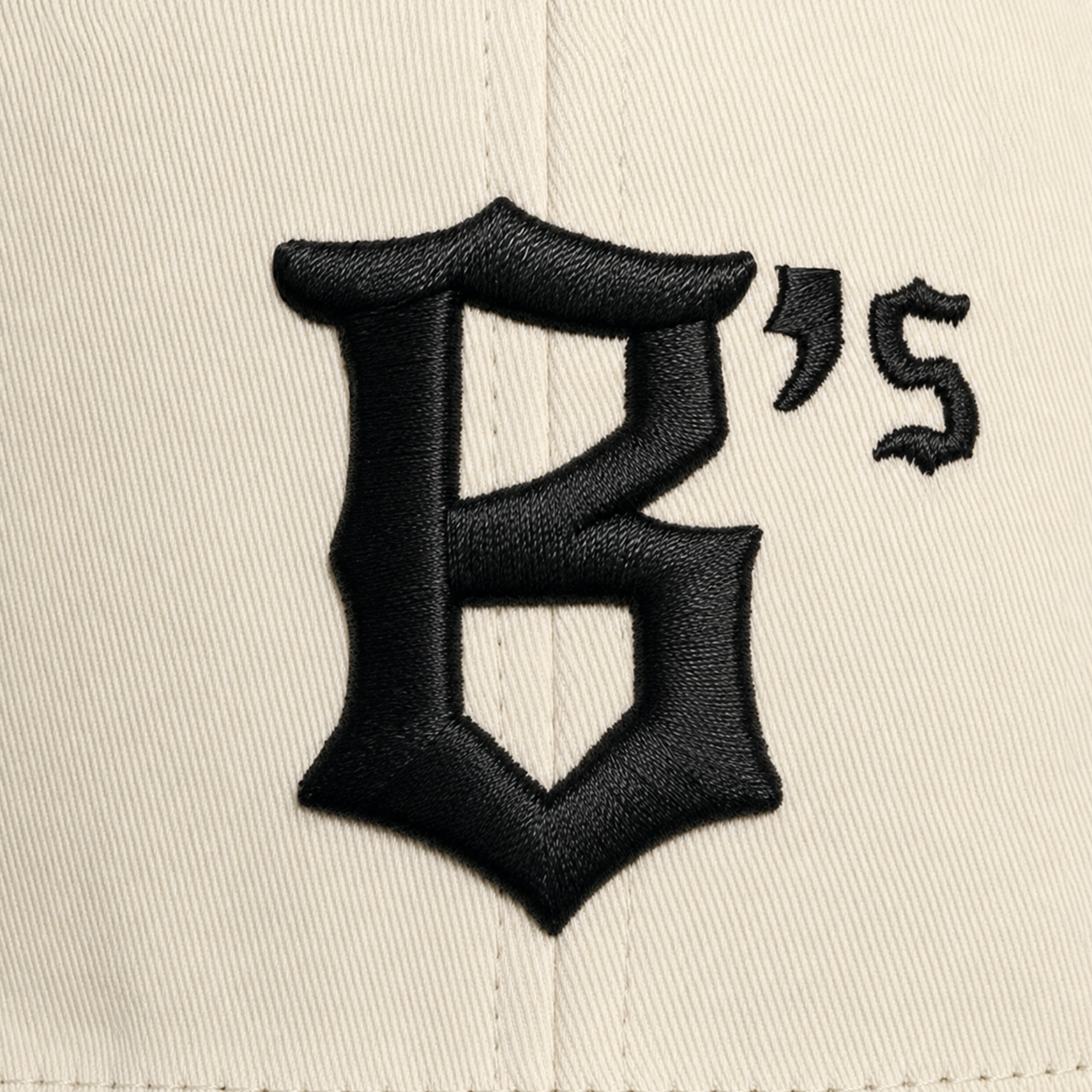 A close-up of the Oakland Peloteros Hat by Oakland Ballers shows beige fabric with prominent, textured stitching and bold, black, embroidered Gothic-style lettering: a large "B" followed by a small apostrophe and "S" in a similar font—distinctive details that evoke the classic style of the Oakland Ballers, an exciting new addition to the Pioneer League.
