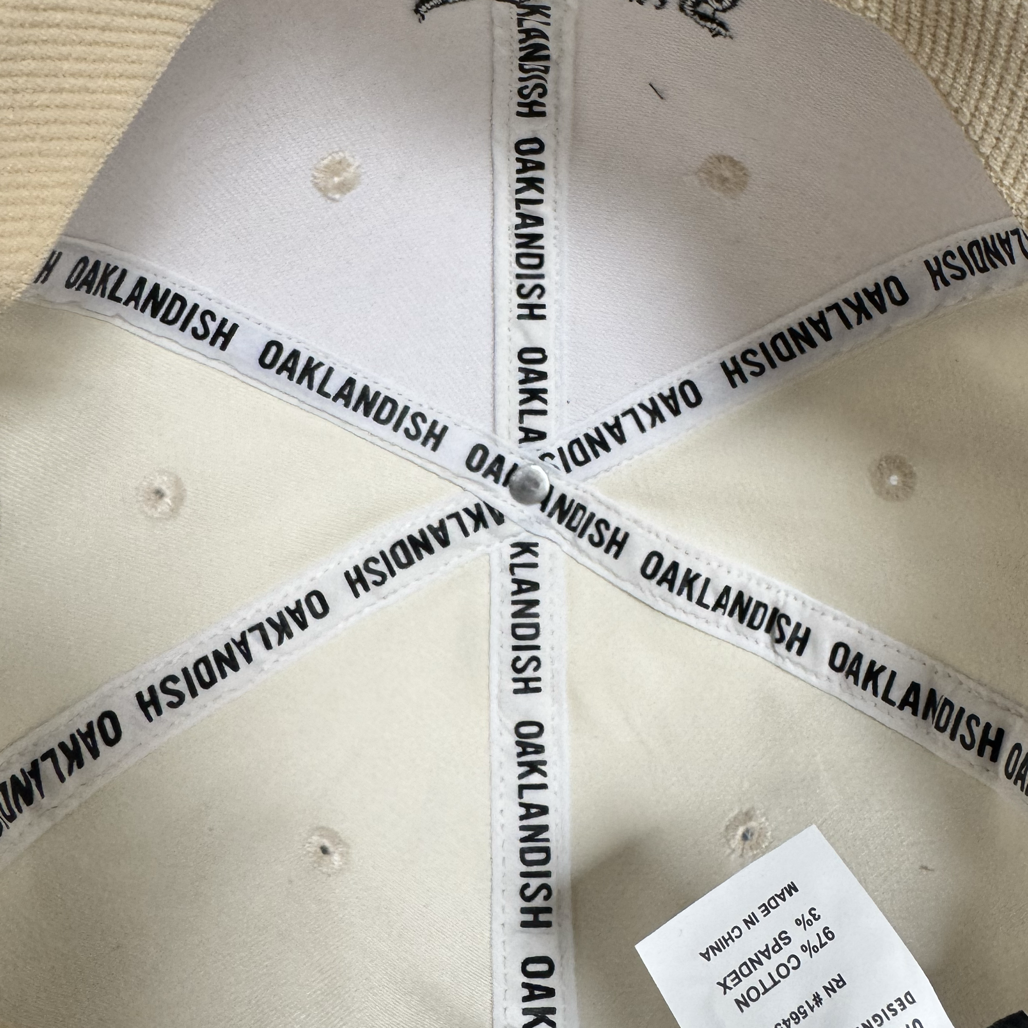 The inside of the Oakland Peloteros Hat by Oakland Ballers reveals white fabric panels joined by white tape featuring repeating black "OAKLANDISH" text. A partially folded white fabric tag, printed with manufacturing and material details, is visible—subtly honoring the rich legacy of Oakland baseball teams and their ties to local culture.