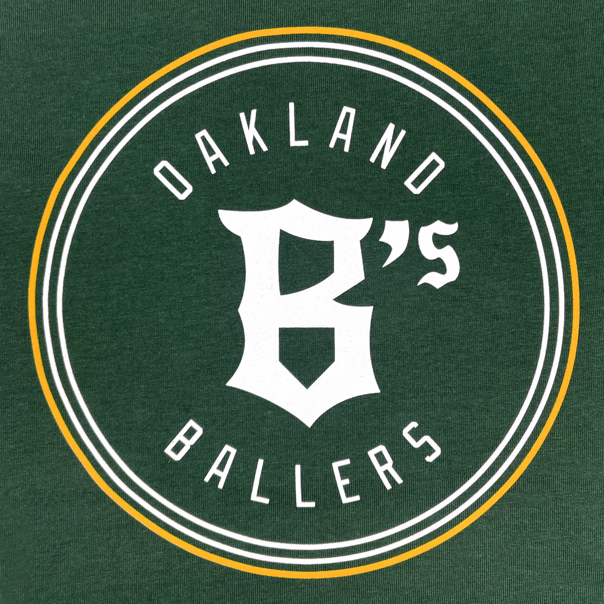 The Infant Oakland Ballers Logo One-Piece showcases the words "Oakland Ballers" in white lettering, encircling a stylized white "B's" in the center. The design, representing a West Coast franchise in the Pioneer League, is set against a forest green background and bordered by a double-lined circular frame in white and gold. This iconic branding is flawlessly incorporated into the one-piece apparel by Oakland Ballers.