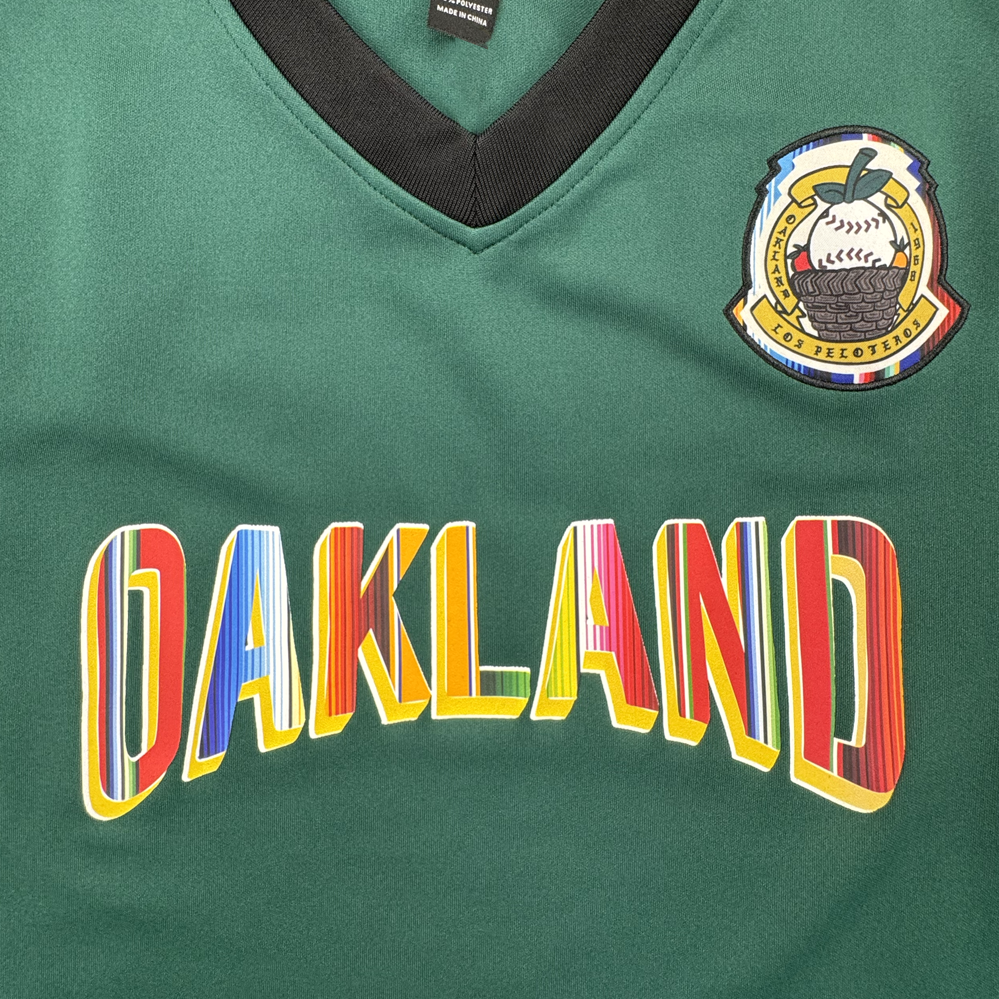 Close-up of a green V-neck jersey with a black collar trim, a multicolor Oakland Peloteros  patch on the upper right chest, and large arched 'OAKLAND' lettering across the front in red, yellow, blue, and white.