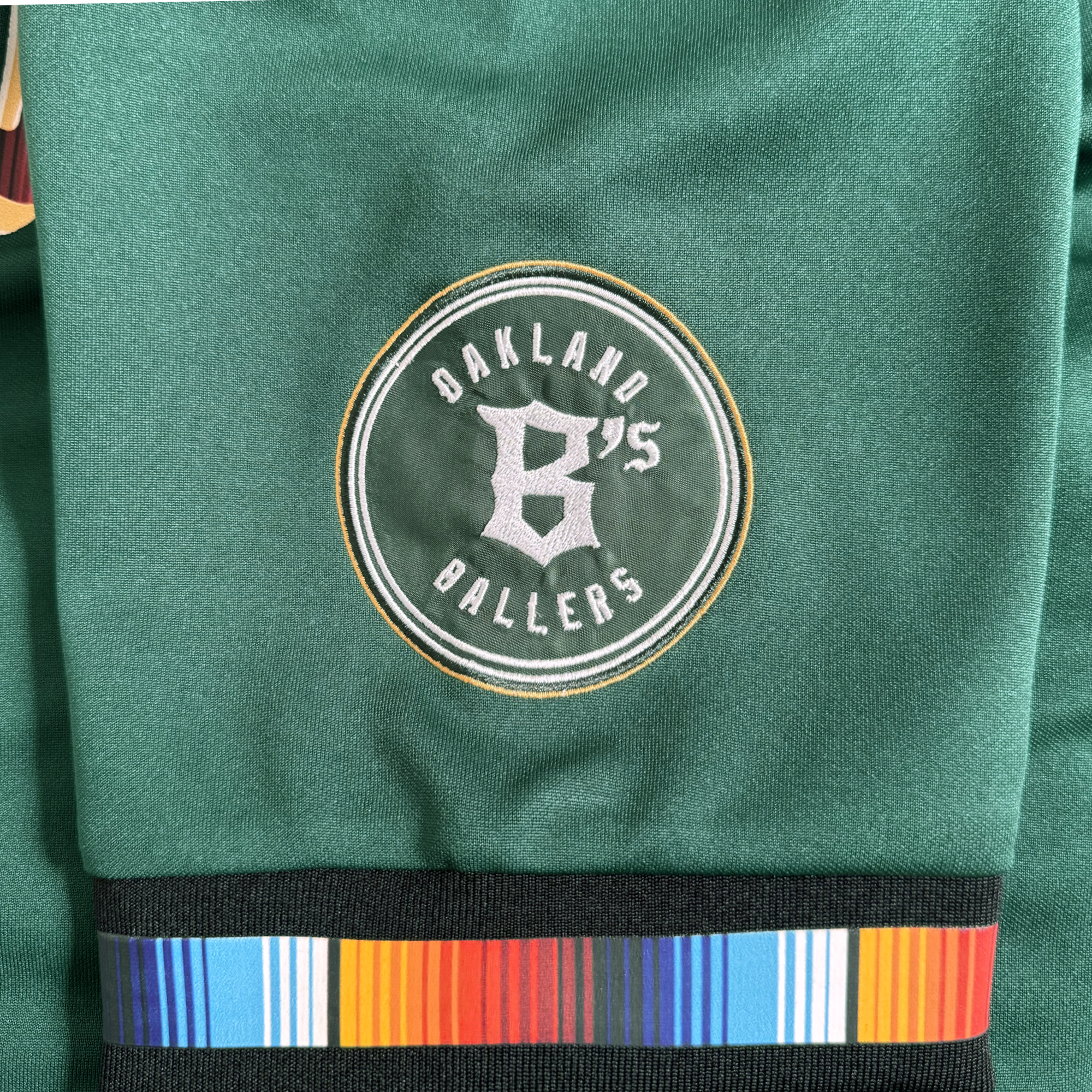 The Oakland Ballers Peloteros Jersey is green and showcases a round patch featuring "Oakland 6's Ballers" with a stylized 6 at the center, honoring the city's newest Pioneer League baseball team. Beneath the patch, a black band displays a vibrant stripe pattern.
