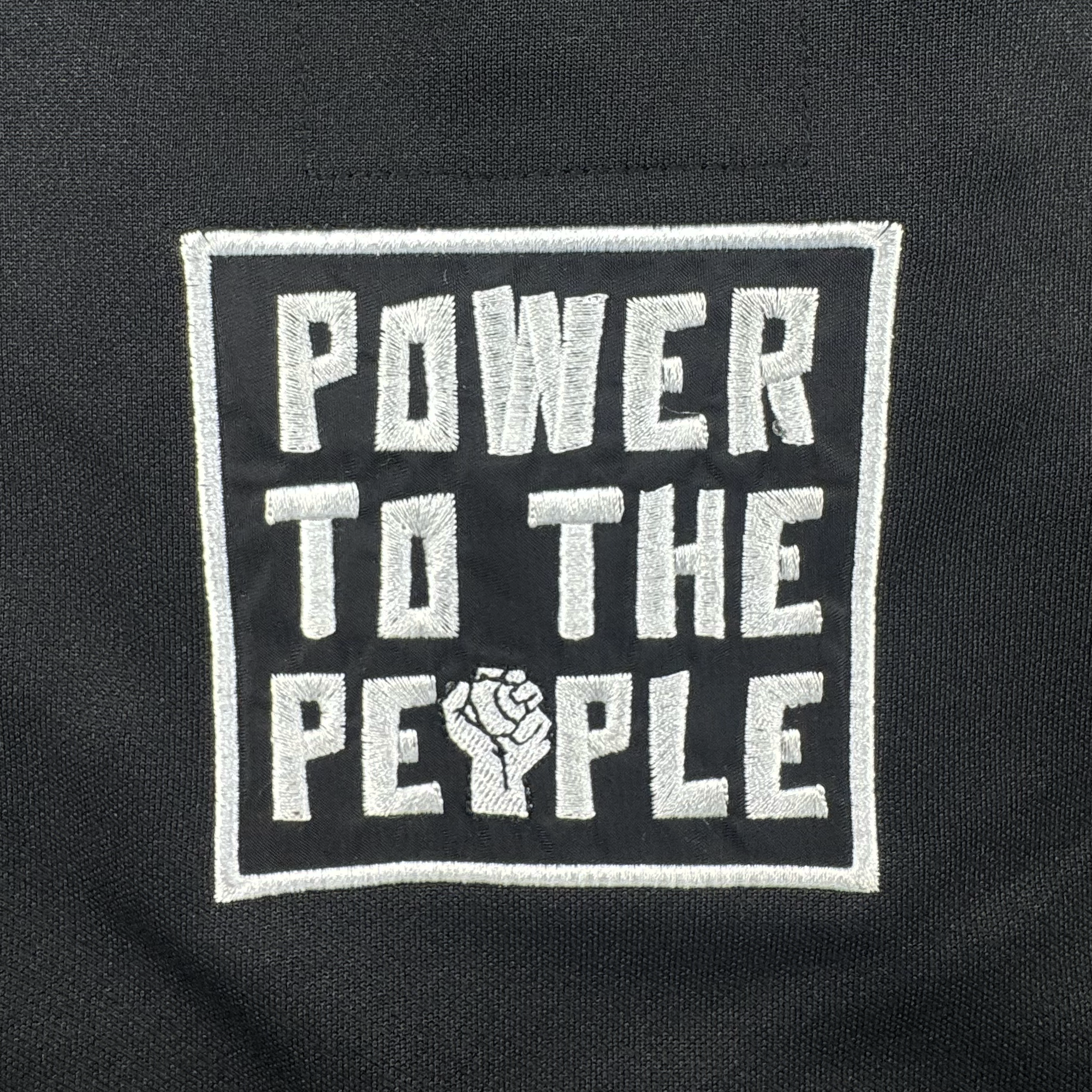 The Oakland Ballers "Stands Up Jersey" features white embroidered text on black fabric reading "POWER TO THE PEOPLE," with a raised fist for the "O" in "PEOPLE"—a bold symbol of solidarity from the new Pioneer League baseball team.