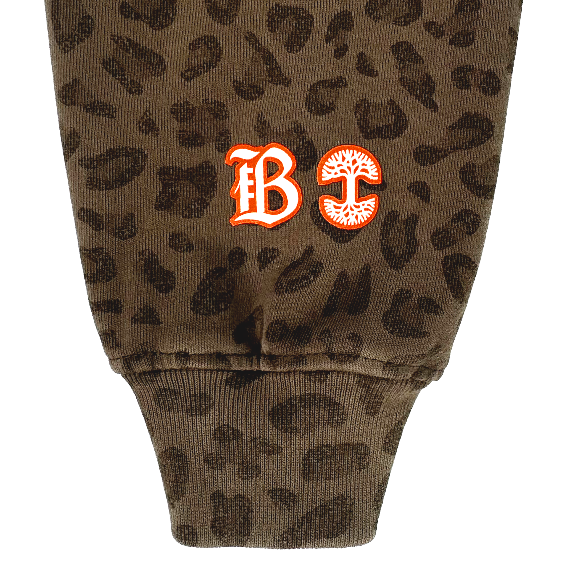 A close-up of the Bay FC Leopard Hoodie shows brown Leopard Camo fabric and a ribbed cuff. Near the cuff are two embroidered designs: a red gothic "B" with white trim, and a red "C" with a stylized mushroom. Brand: Bay FC.