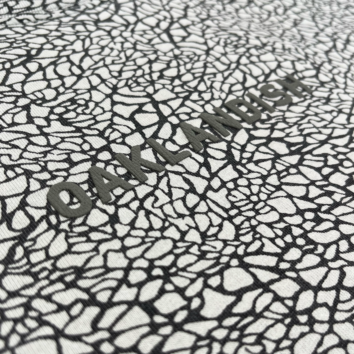 Close-up of the Oaklandish Classic Logo Tee’s textured surface, featuring an irregular, black-and-white cracked pattern. “OAKLANDISH” is prominently embossed across the center in bold, uppercase letters with a distinctive puff print technique that partially merges with the intricate background—showcasing the signature style and heavyweight cotton of this classic fit tee from Oaklandish.