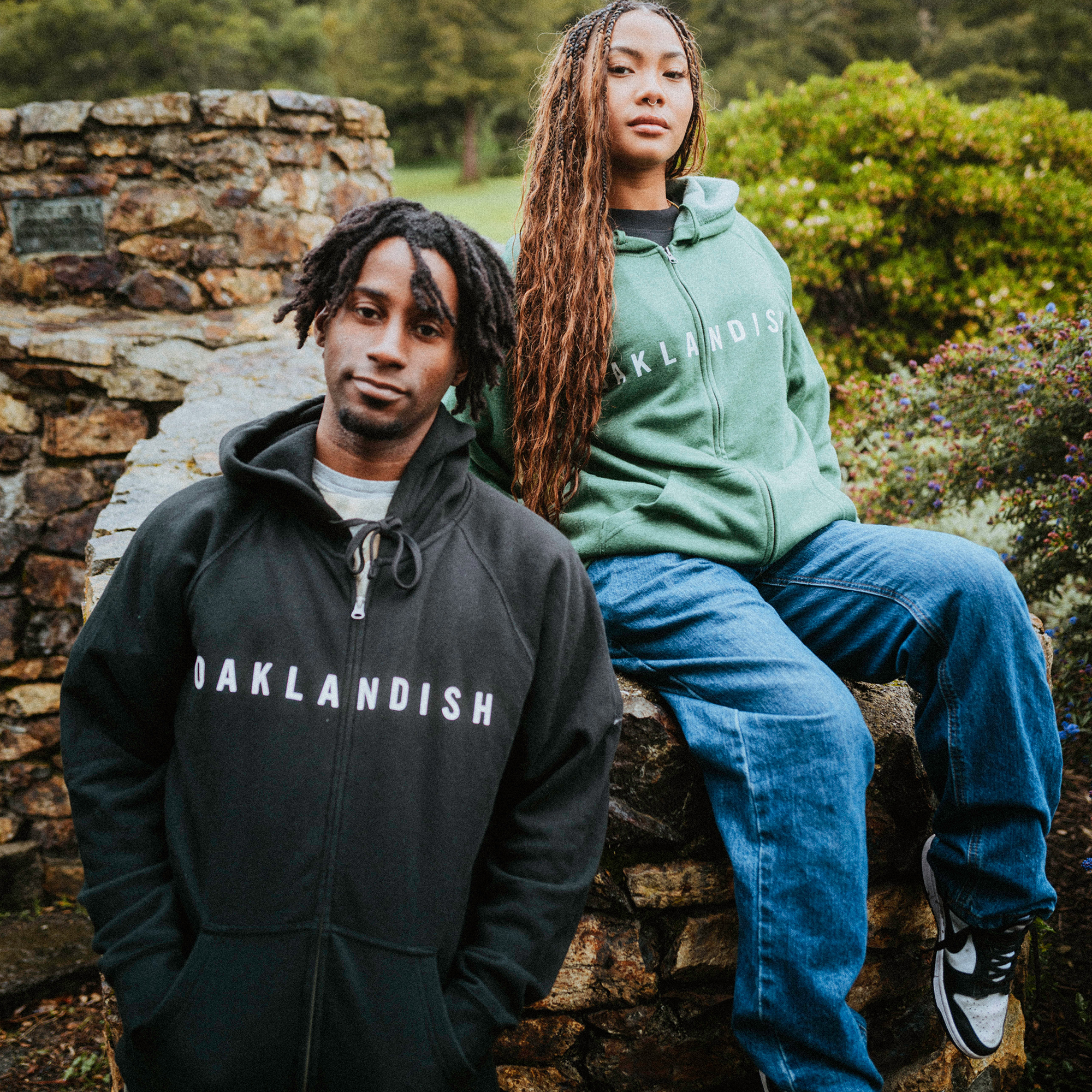 A person in a black Oaklandish hoodie with unisex fit stands with hands in pockets, next to another in a green Oaklandish Classic Zip Hoodie. The seated individual, with long braided hair, jeans, and sneakers, enjoys the outdoors amid greenery and stone structures.