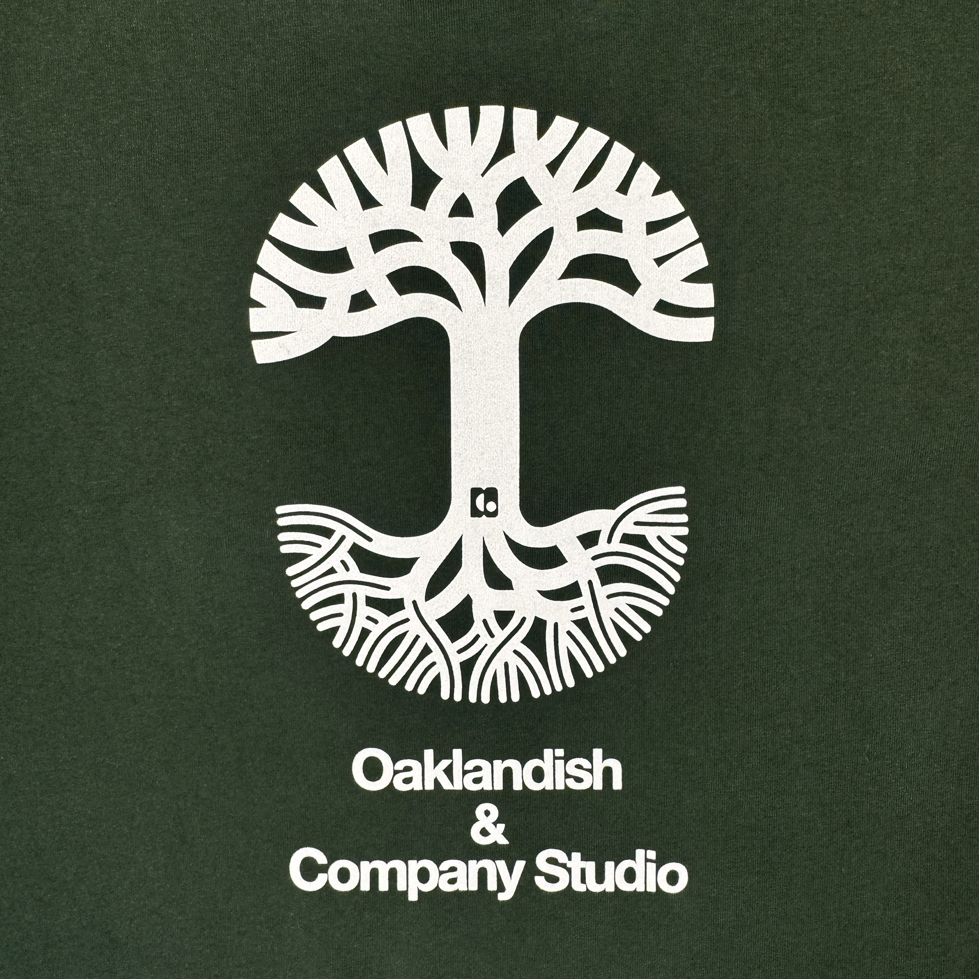 A white tree with stylized branches and roots creates a circular motif against a dark green background, echoing Oaklandish’s iconic design aesthetic. Beneath the tree, crisp white lettering spells out: “Oaklandish & Company Studio.” This clean, modern style serves as the perfect backdrop for The East Bay Tee—a limited-edition release from Oaklandish in collaboration with Toro y Moi’s Chaz Bear.