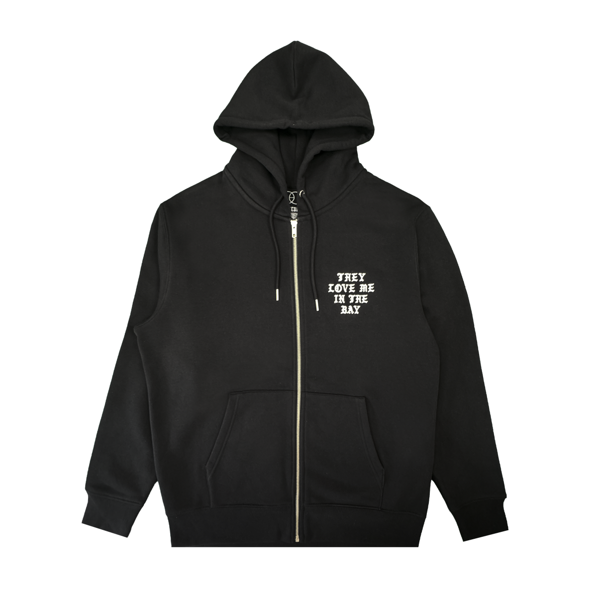 A black They Love Me Zip By Doc hoodie from By DOC, made with heavyweight fleece and designed with front pockets and a hood. The left chest features white, all-caps text that reads “THEY LOVE ME IN THE BAY.” The hoodie is displayed flat on a white background.
