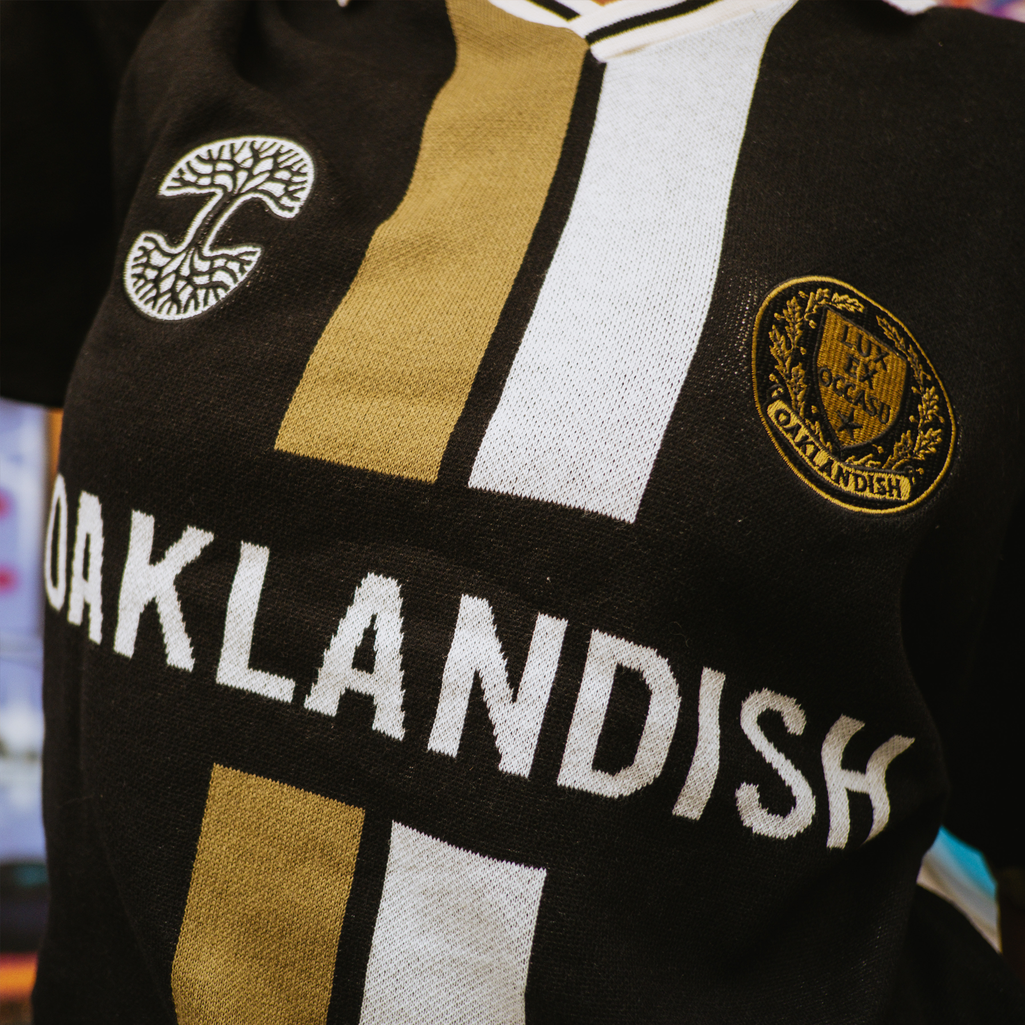A close-up of the Oaklandish Ease Jersey, a men's classic fit knit collar jersey in black, gold, and white, featuring "OAKLANDISH" in bold letters, a white tree emblem on the left, and a gold oval patch with the tree design on the right.