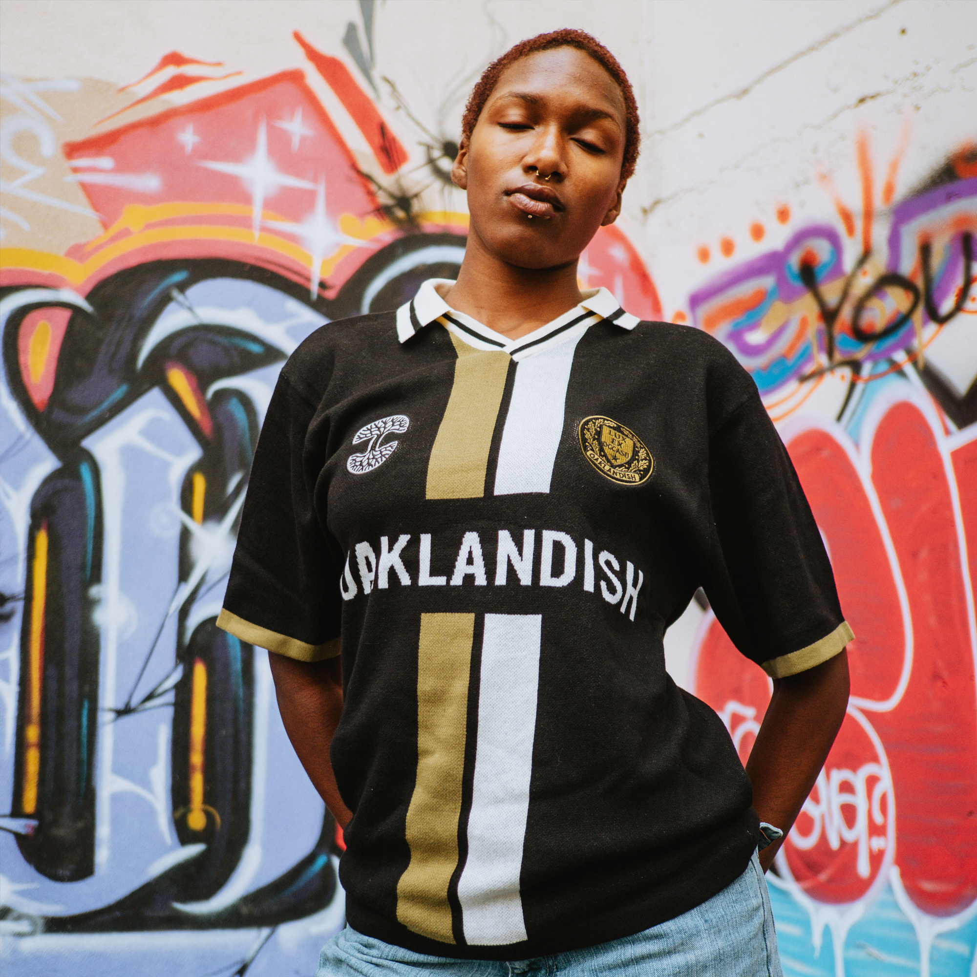 A person with short hair and a nose piercing stands confidently before a colorful graffiti wall, wearing an Oaklandish Ease Jersey in black and gold and light blue jeans, looking slightly down at the camera.