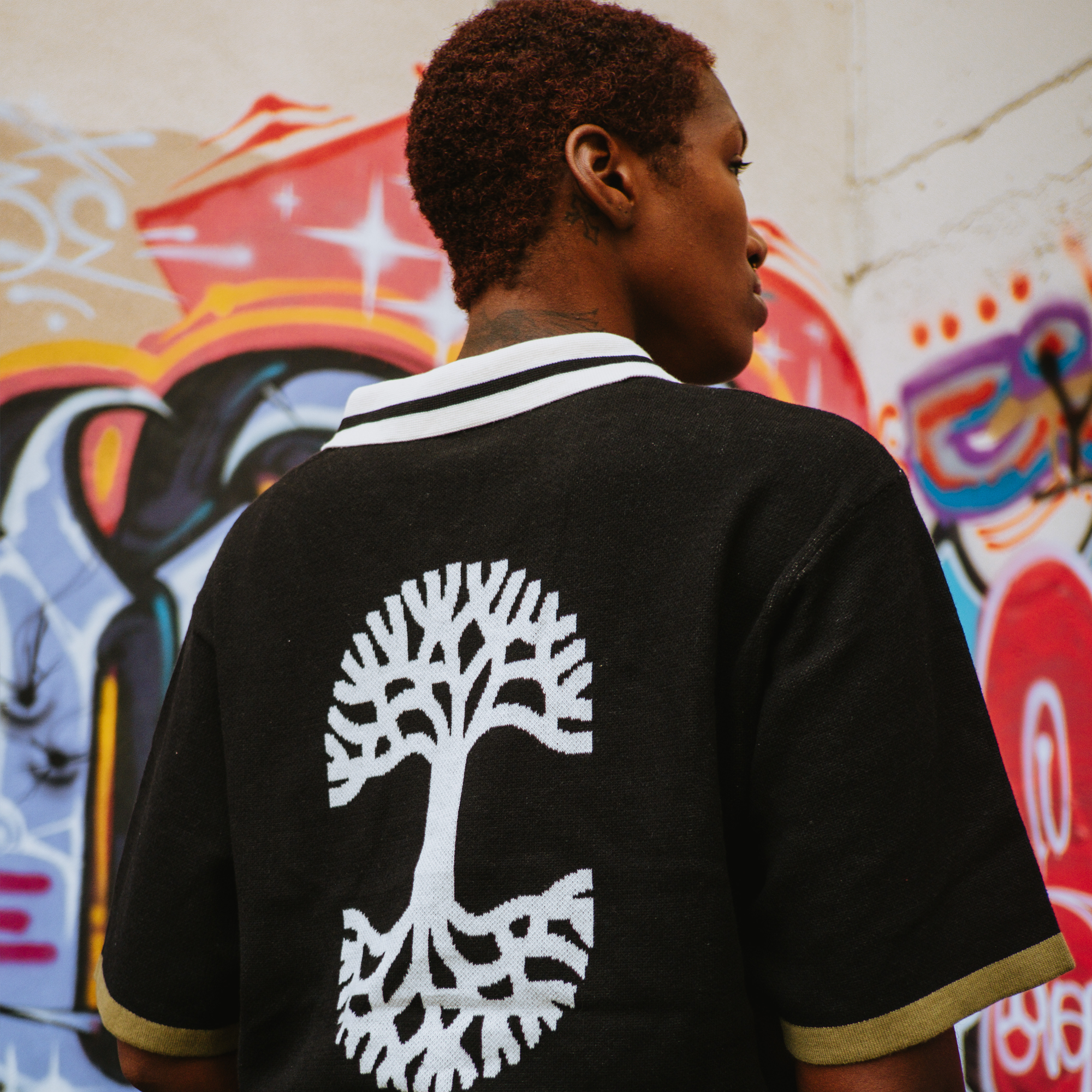 A person with short curly hair stands sideways, showing the back of an Oaklandish Ease Jersey with a large white tree graphic. The classic fit black jersey is set against a vibrant graffiti wall in reds, blues, and oranges.