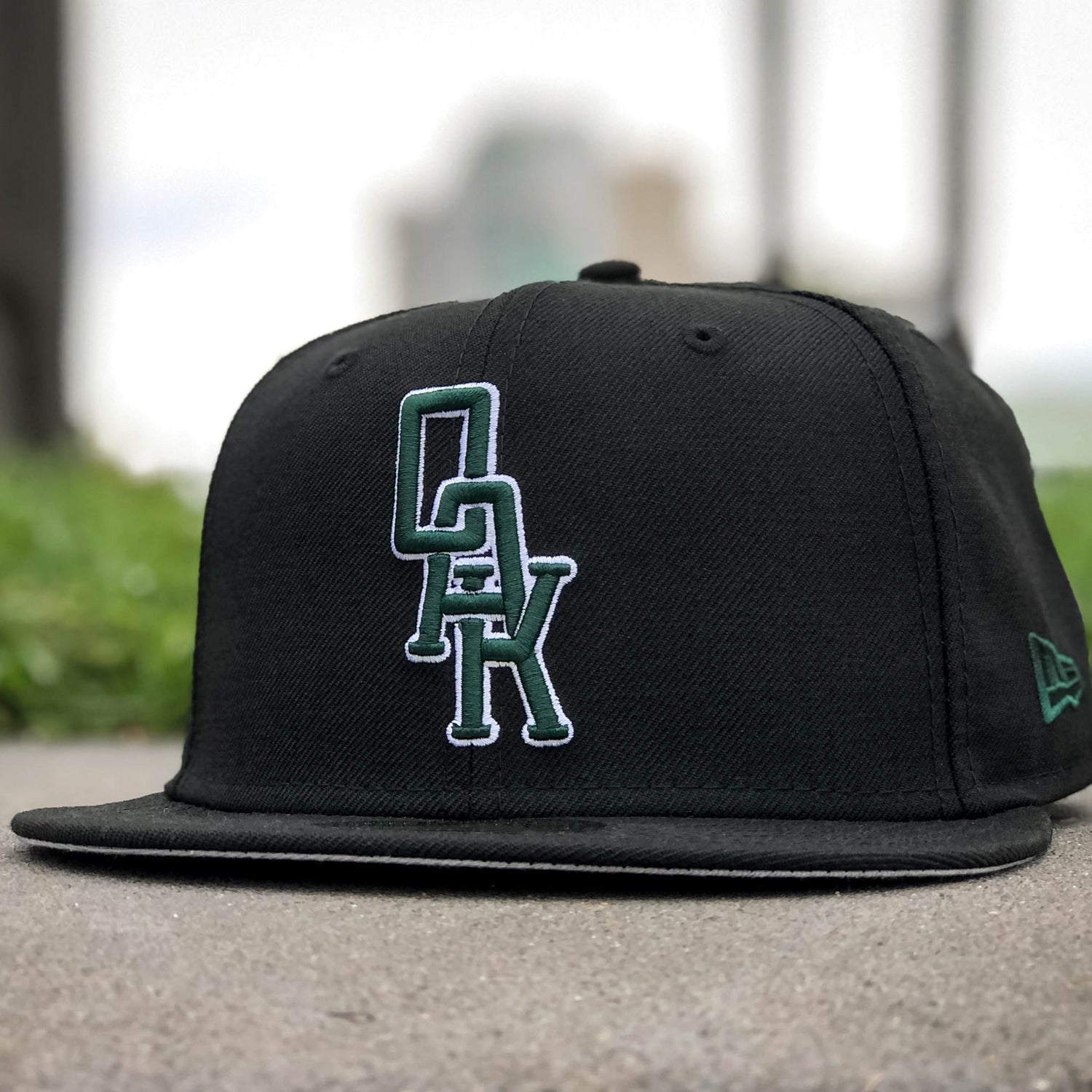 A black New Era OAK 59FIFTY Fitted Cap by Other sits on concrete. The front features the distinctive OAK logo, with bold white letters outlined in green stacked vertically. In the background, green grass and a blurred cityscape can be seen, while the signature New Era logo is stitched on the side to signify its high quality.