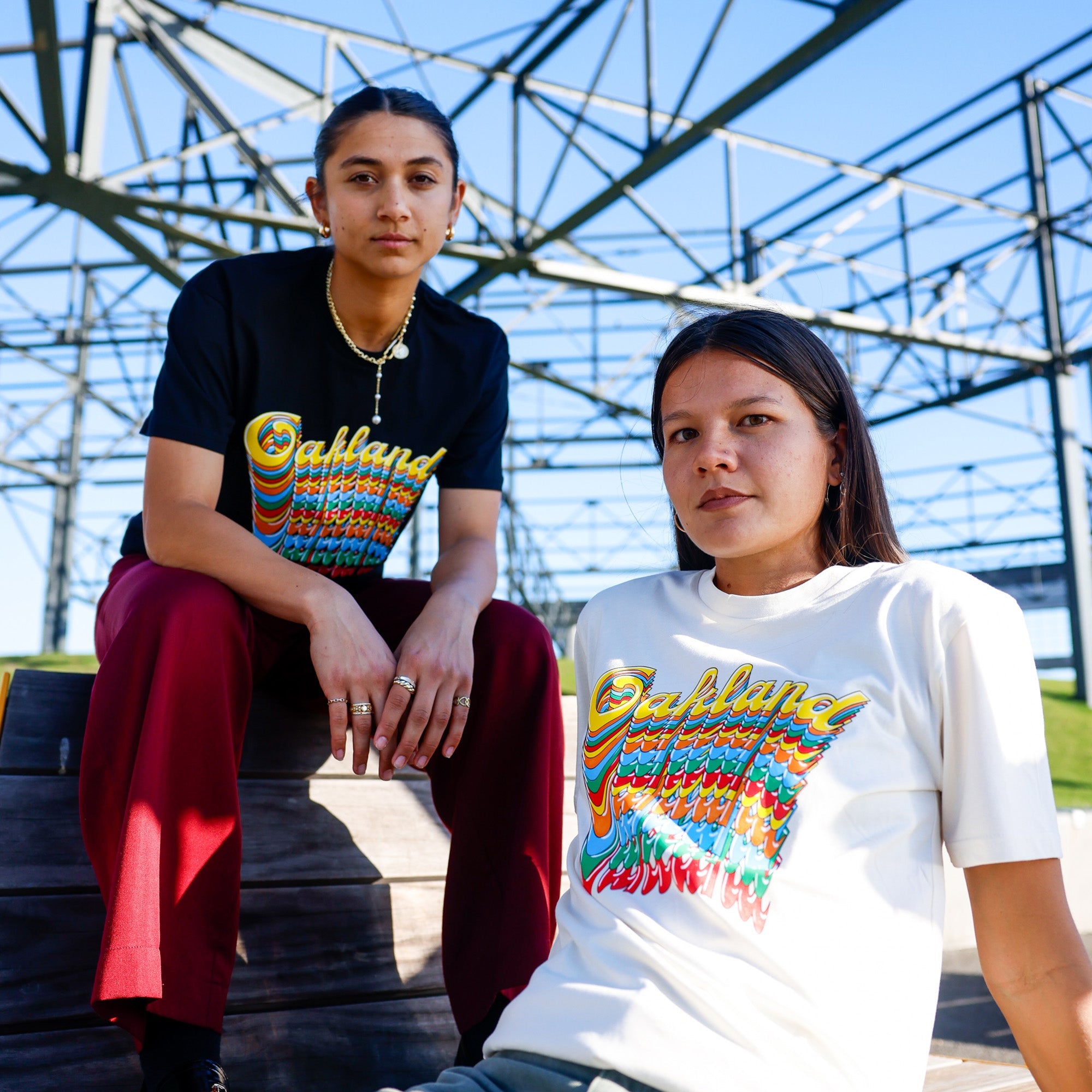 Two individuals pose outdoors under a structure with geometric metal beams. One sits on a wooden platform wearing a "Oakland Roots SC Funk Tee" and red pants. The other person, seated lower, wears a white "Oakland" graphic T-shirt. The sky is clear and blue.